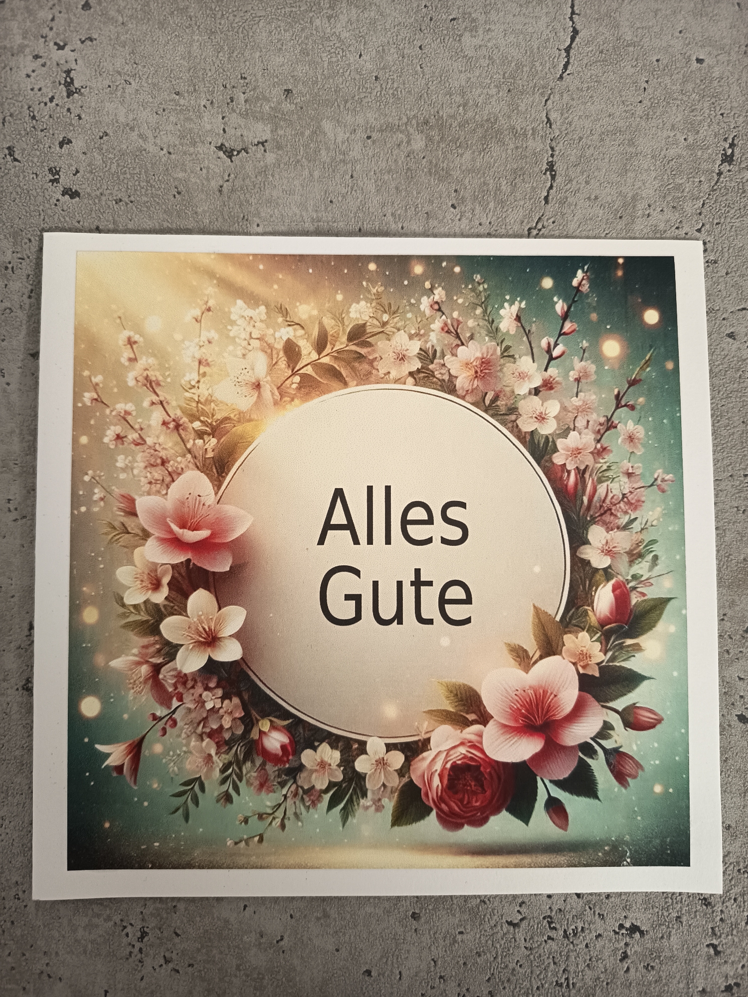 The image shows a greeting card on a grey surface. The card has a floral design surrounding a cream-colored circle. Inside the circle, in black letters, is the German phrase "Alles Gute," which translates to "All the best" or "Best wishes." The flowers are pink and white, with some greenery and twinkling light effects in the background. The card appears to be printed on glossy paper.
The image does not contain information about the location in the city of Wangen bei Olten, Switzerland.