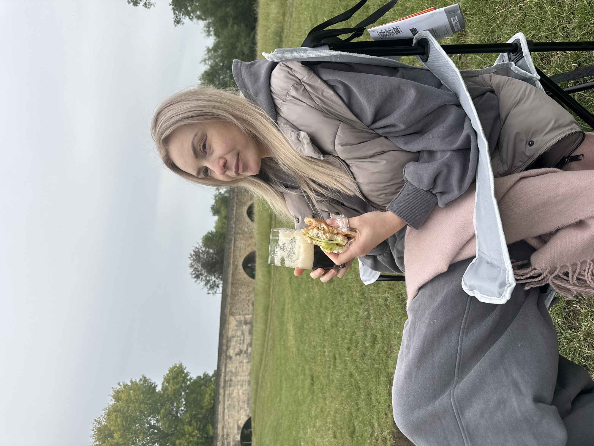 A blond woman, with fair skin and light facial freckles, is seated in a light grey folding camping chair on a grassy field under an overcast sky. She is looking towards the left side of the frame with a neutral expression. Her hair is parted slightly to the left, falling over her shoulders. She wears a grey hooded sweatshirt layered under a taupe-colored quilted sleeveless vest, and dark grey sweatpants. A light pink fringed blanket or scarf is draped over her lap.
In her right hand, she holds a clear glass mug filled with a dark, frothy beverage, likely a stout or similar dark beer. In her left hand, she holds a half-eaten sandwich with green leafy contents and what appears to be chicken or another filling.
In the mid-ground, behind the woman, a long stone bridge or viaduct with multiple arches spans across the scene. The bridge is made of light-colored stone, and parts of it are partially obscured by trees. The foreground and mid-ground consist of green grass, showing some worn paths or tire tracks. Various trees with green and darker foliage are scattered throughout the background. A white tag with a black QR code and printed text (partially visible: "HHN 50970-2507" and "1Z") hangs from the right armrest of the chair. The location is an outdoor rural or park setting, specifically in the city of Unknown, Unknown.
