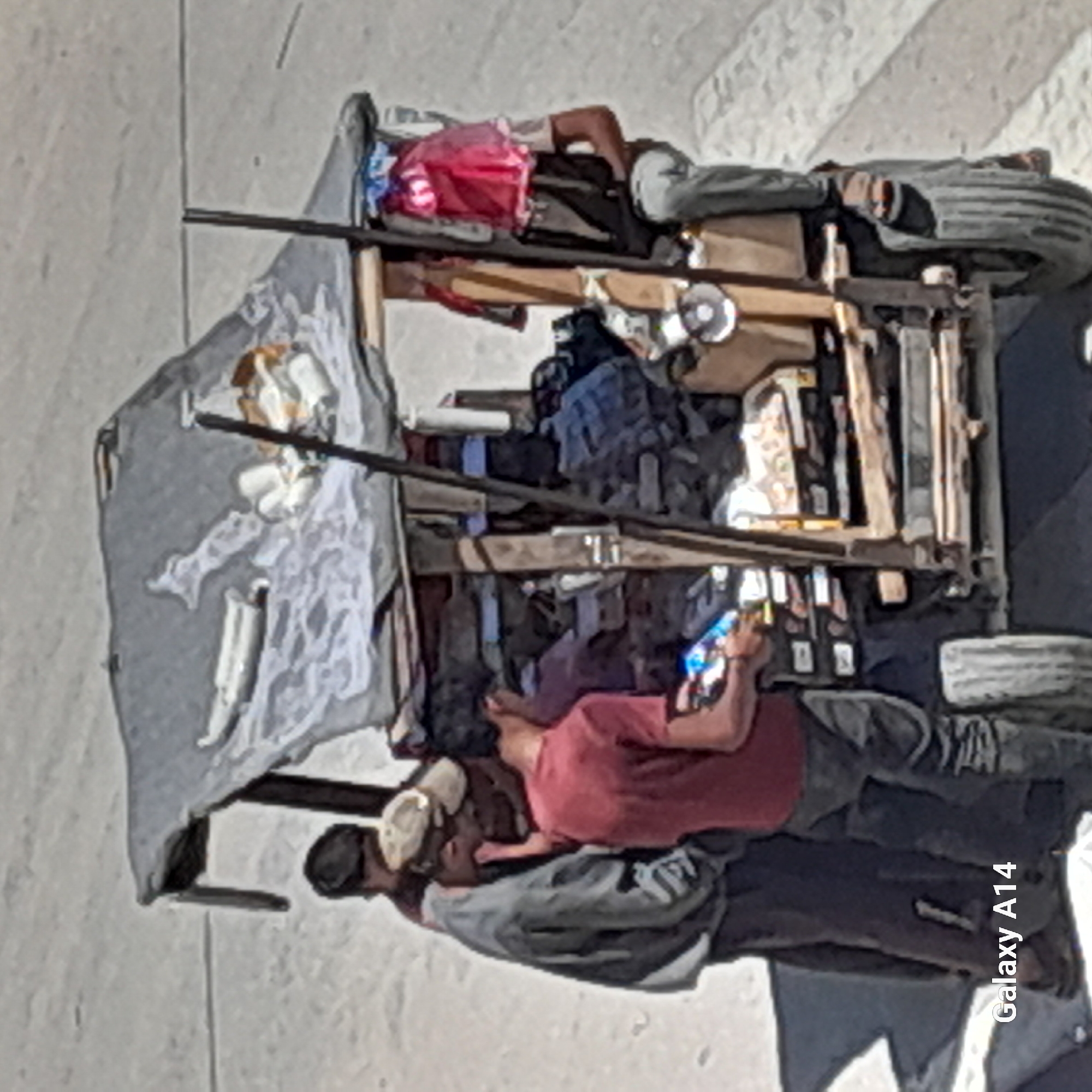 In Khan Yunis, Palestinian Territories, a makeshift market stall, likely a cart, is set up. The stall has a dark, tarp-like covering stretched over a wooden frame, providing shade. Several people are gathered around the stall. One person, wearing a red shirt and dark pants, is holding a colorful packaged item, possibly food or a drink. Another person, in a grey shirt and dark pants, is leaning forward, looking towards the stall. A third individual, wearing a beige cap, is partially obscured. The stall itself appears to be laden with various items, including what seem to be packaged goods and possibly fabric. The ground surface is paved, suggesting an urban or semi-urban environment. The overall impression is a bustling scene at a street vendor's setup.