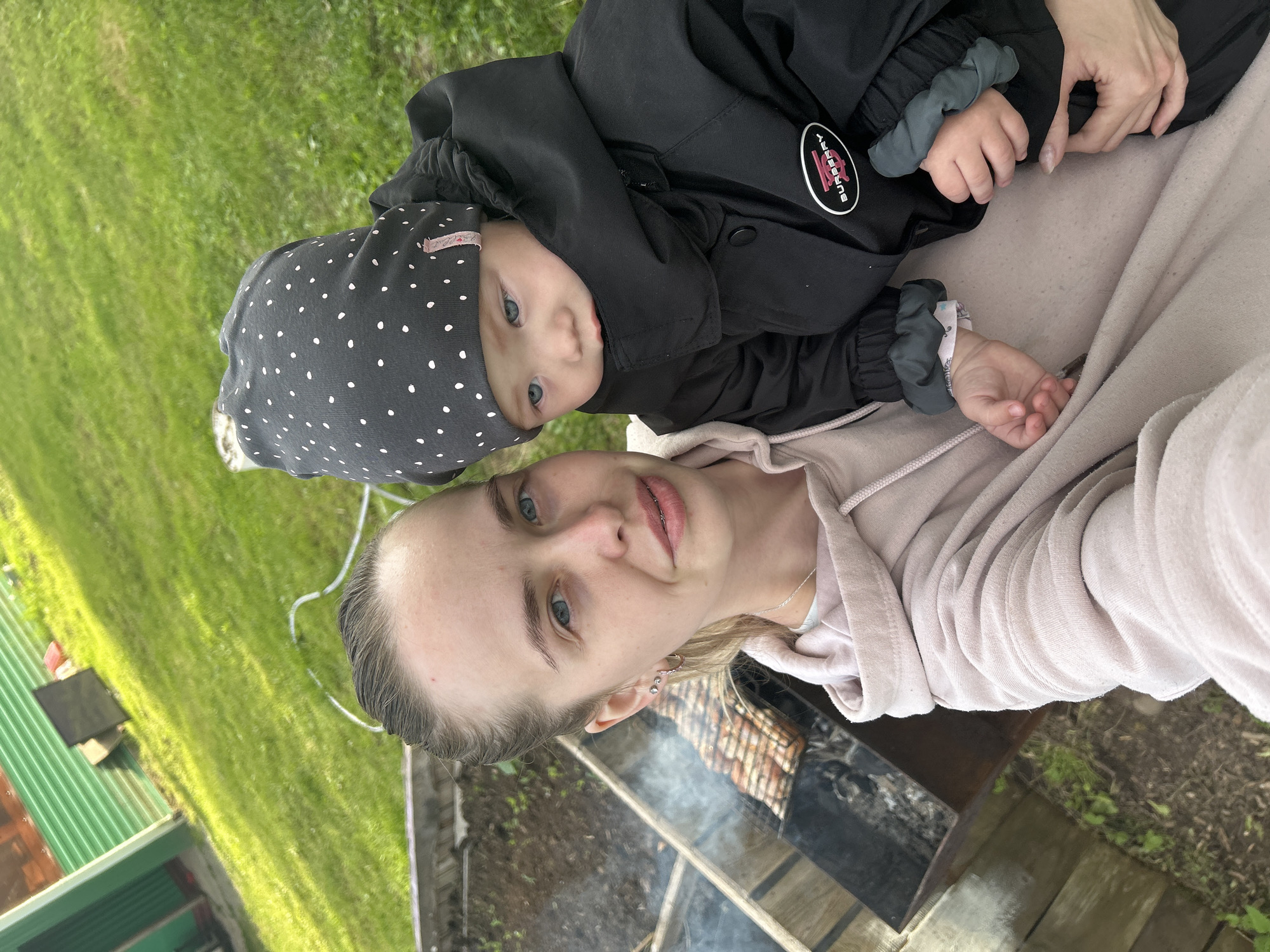 In Азинский, Russia, a woman with blonde hair pulled back in a ponytail is taking a selfie outdoors. She is wearing a light pink hoodie and holding a baby. The baby is wearing a black puffer jacket with a dark gray polka dot beanie. Behind them, a barbecue grill with food cooking over coals is visible. The background consists of a green corrugated metal fence, a grassy yard, and some dirt. The lighting suggests it is daytime.