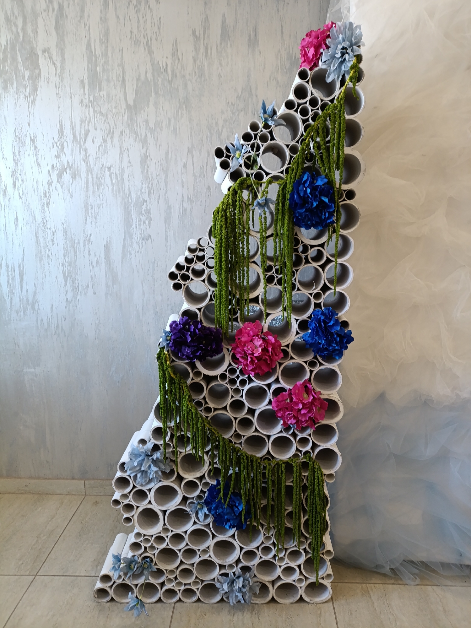The image presents a handcrafted decorative structure, presumably an art installation or backdrop. It is constructed from an arrangement of white PVC pipes of varying diameters, arranged in a stacked, uneven vertical formation. The structure is adorned with artificial flowers in shades of pink, blue, and purple. Trailing green garlands are draped across the structure, adding a touch of natural element.
The backdrop is a textured, light gray wall. The flooring appears to be made of large, light-colored tiles. A gauzy, white fabric is partially visible on the right side of the frame.
Without external location references or landmarks within the scene, the statement indicating this is located in Chaikovsky, Russia, cannot be verified from this image alone.