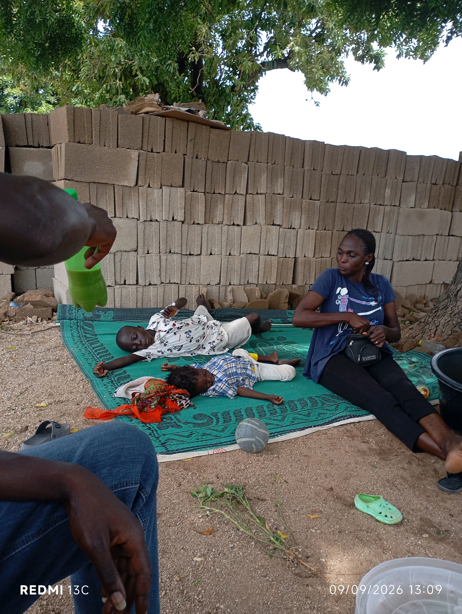 In Shendam, Nigeria, two young children lie asleep on a green mat placed on the ground. One child is dressed in a white shirt with a Mickey Mouse pattern and white pants, while the other wears a blue and white plaid shirt. A woman in a blue t-shirt and black pants sits next to them, looking away and holding a small black bag. A hand holding a green bottle of liquid is visible in the foreground, as are several objects including a colorful cloth, a gray ball, a green sandal, a bucket, and some plant debris. A wall constructed from concrete blocks serves as the backdrop, with trees and foliage overhead. The scene appears to be outdoors, possibly in a yard or a makeshift gathering space.
