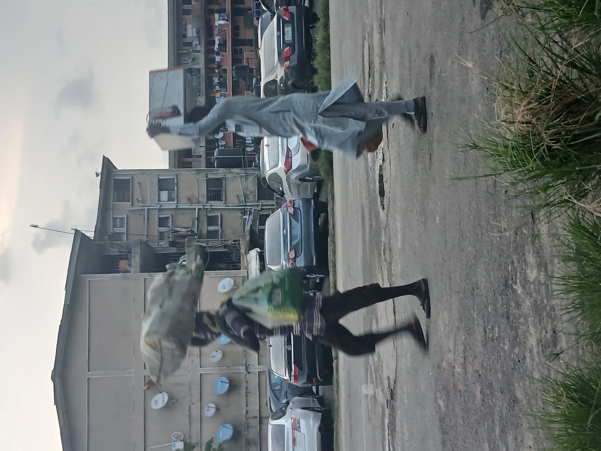 The image depicts an urban scene in Lagos, Nigeria. Two people are walking along a paved road lined with parked cars, with apartment buildings in the background. One person is carrying a large sack over their shoulder, while the other is carrying a box. The surroundings include buildings with satellite dishes attached and patches of grass.