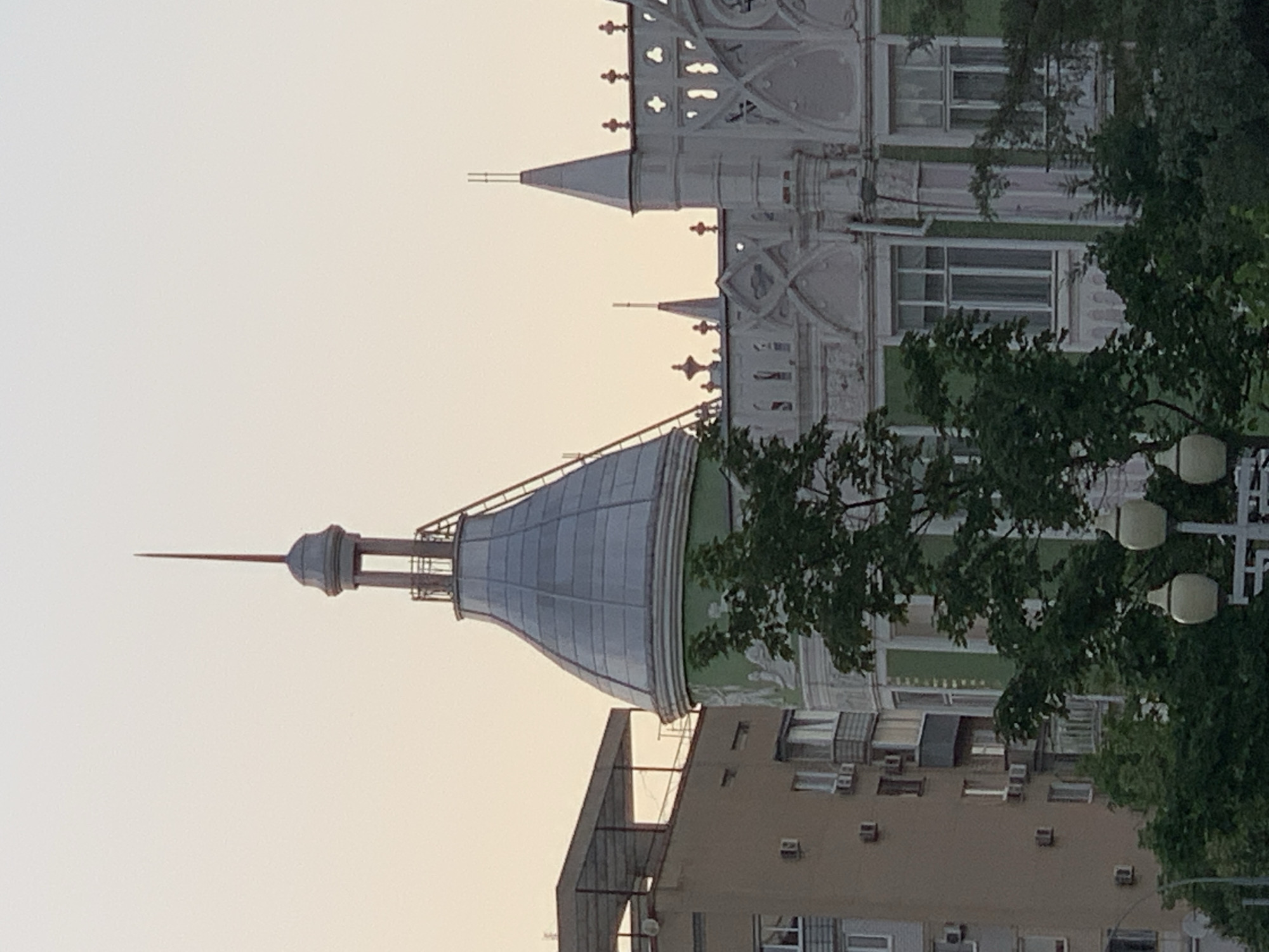 The image captures a portion of an urban skyline at dusk or dawn. Dominating the scene is a tall, conical turret with a silver, segmented roof, topped by a thin, pointed spire. A ladder is attached to the side of the turret, ascending towards the peak. To the left of the turret, a modern, multi-story apartment building with numerous windows and air conditioning units is visible. The building has a flat, extended roof structure. To the right of the turret, a white, ornate building with gothic-inspired architectural elements stands out, featuring intricate detailing, pointed arches, and decorative pinnacles. Green trees and foliage obscure the lower portions of the buildings, with a street lamp visible in the foreground. The sky is a pale orange and yellow gradient, indicating the time of day.