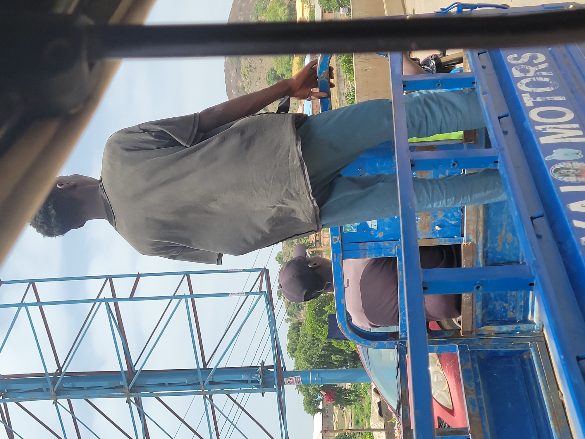 From within a vehicle, a man in a gray shirt and teal pants drives a blue tricycle through Suleja, Nigeria. Another man, wearing a black cap and brown shirt, rides in the back. The vehicle is branded with "JAIJO MOTORS." In the background, a hill and a billboard structure can be seen. Other vehicles are visible in the periphery.