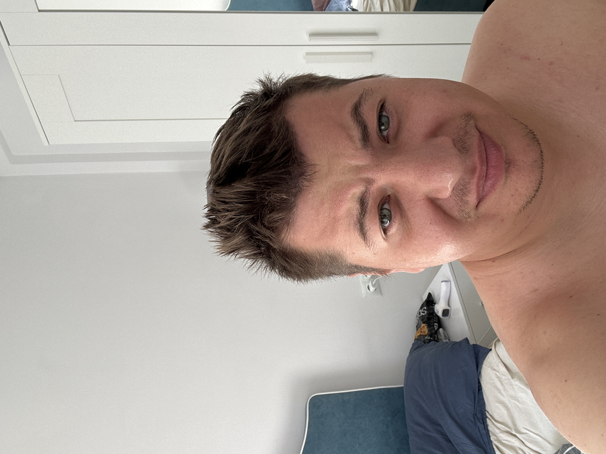 The image is a selfie of a man taken indoors, likely in a bedroom. The man has short, messy brown hair, green eyes, and is shirtless. He has a slight stubble on his chin and upper lip. He is looking directly at the camera with a neutral expression. In the background, there is a white closet with two doors and a mirror on the right side. To the left of the closet, there is a white wall and part of a blue headboard of a bed. On a nightstand next to the bed, there is a dark blue pillow, a bag of chips, and a white digital thermometer. The scene appears to be in a domestic setting. The image does not provide any specific clues to its location within Benimàmet-beniferri, Spain.