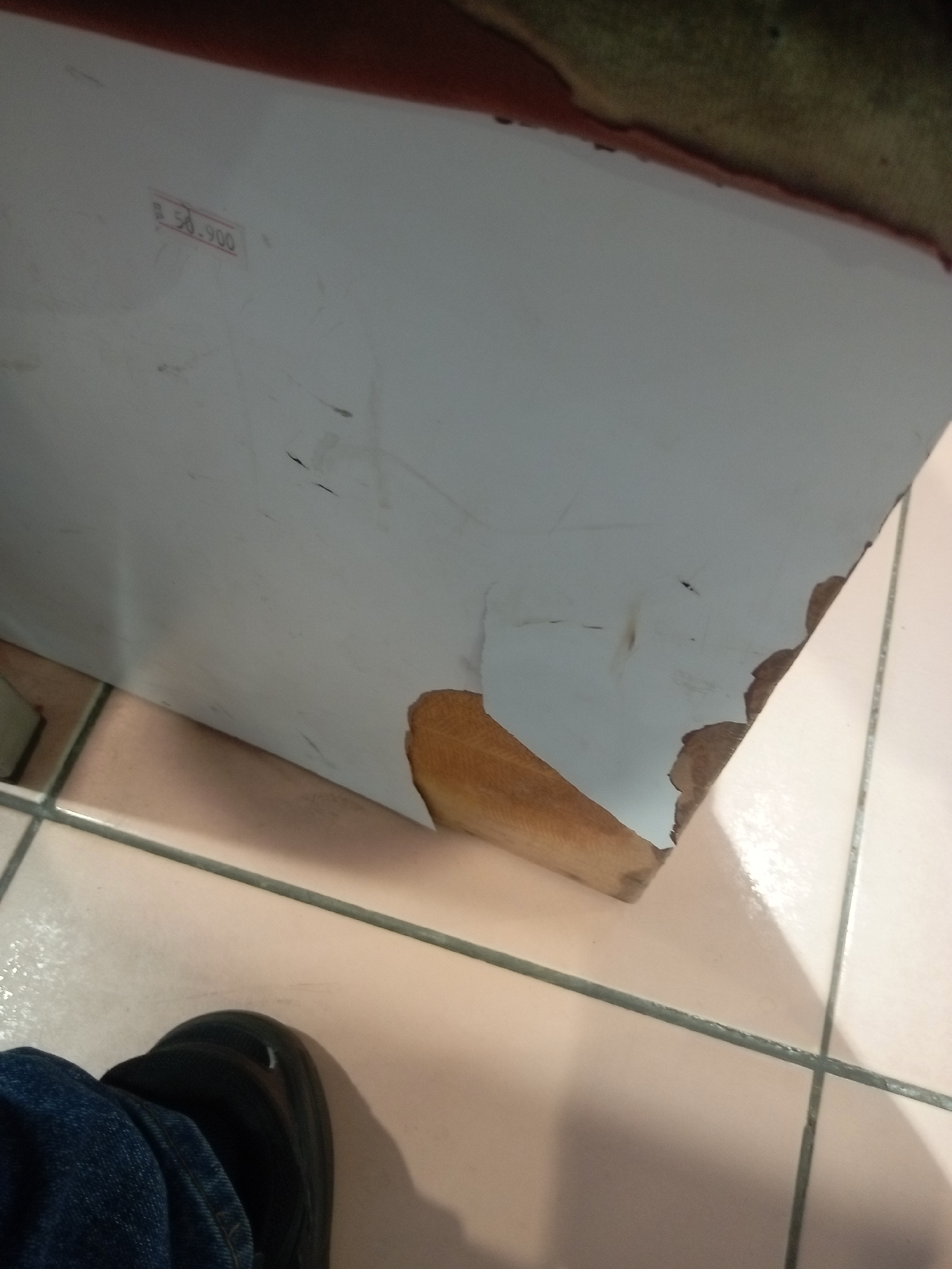 A price tag is visible on the corner of a white surface, reading "B 50.900". The white surface appears to be part of a piece of furniture with damaged edges, exposing a wooden interior. The furniture rests on a tiled floor with light pink tiles and dark grout lines. In the lower left corner, a person's feet are visible, wearing blue jeans and black shoes. The overall scene suggests an indoor environment, possibly a retail or storage area in Batam, Indonesia, given the currency and common tiling style in the region.