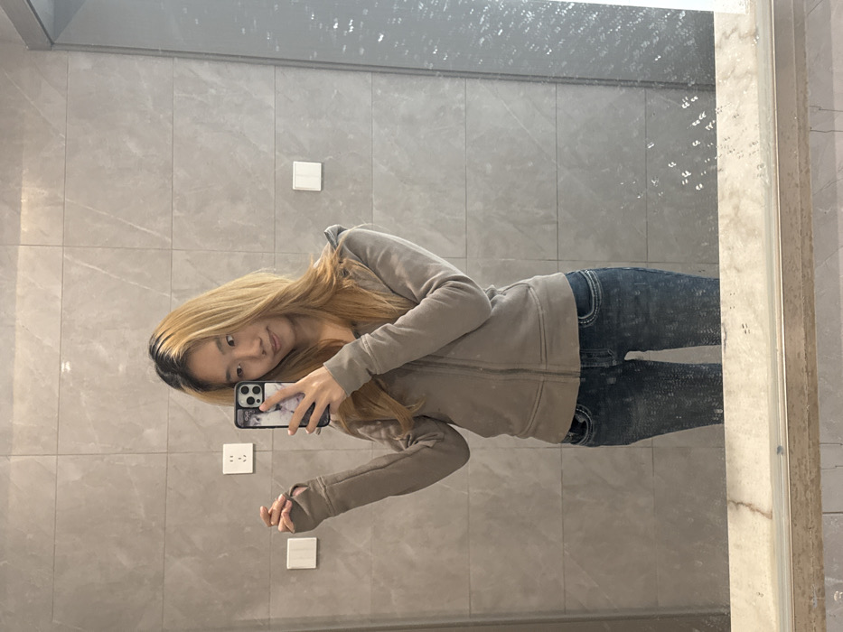 An East Asian female is depicted taking a mirror selfie in an indoor setting. She has long, light brown hair with darker roots and a slight smile. She wears a light brown/taupe zippered hoodie with thumbholes on the sleeves and dark blue distressed jeans. Her right hand holds a smartphone with a patterned case, angled towards the mirror to capture the reflection. Her left arm is bent, with her hand slightly raised, showcasing the thumbhole. The background consists of a wall covered in large, rectangular light gray tiles with subtle veining. Two white electrical outlets/switches are visible on the left side of the wall, and one similar white switch is on the right. Below the mirror, a light-colored, possibly marble, counter or shelf is present. The mirror itself exhibits some smudges and splatters, particularly on the right side. While the prompt specifies Nan'an District, China, the image contains no specific visual identifiers for this location; the environment appears to be a modern interior space, potentially a bathroom or dressing area.