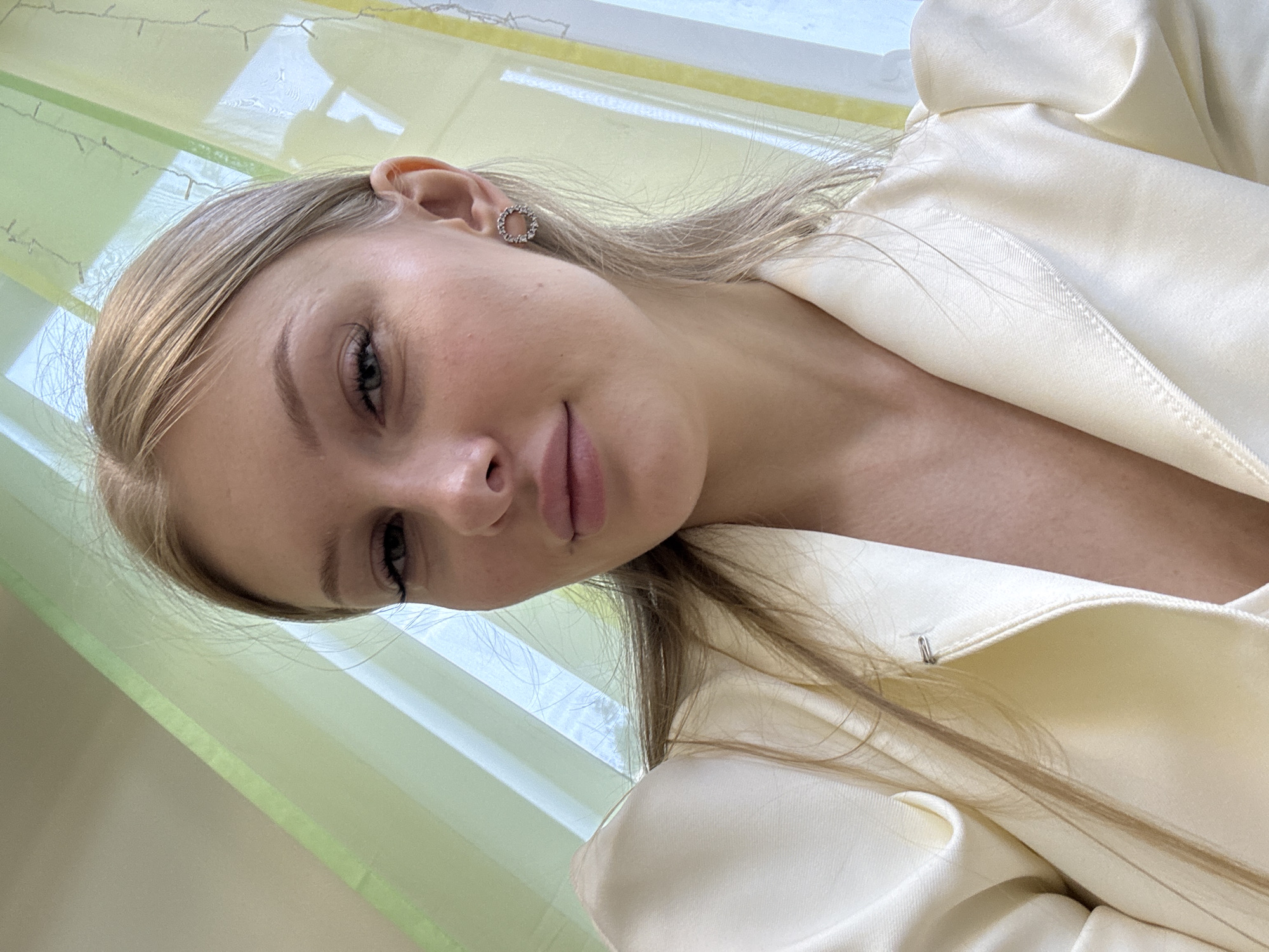This is a portrait photo of a young Caucasian woman with blonde hair, taken indoors. She is wearing a cream-colored blazer with puffed sleeves and a circular, sparkling earring in her left ear. The background consists of green and yellow vertical blinds, and a window showing an outdoor scene, possibly with trees. The lighting is natural, coming from the window. The image appears to be taken at a slightly high angle, looking down at the woman. There is no information about the location in the city of Perm, Russia, in the provided image.