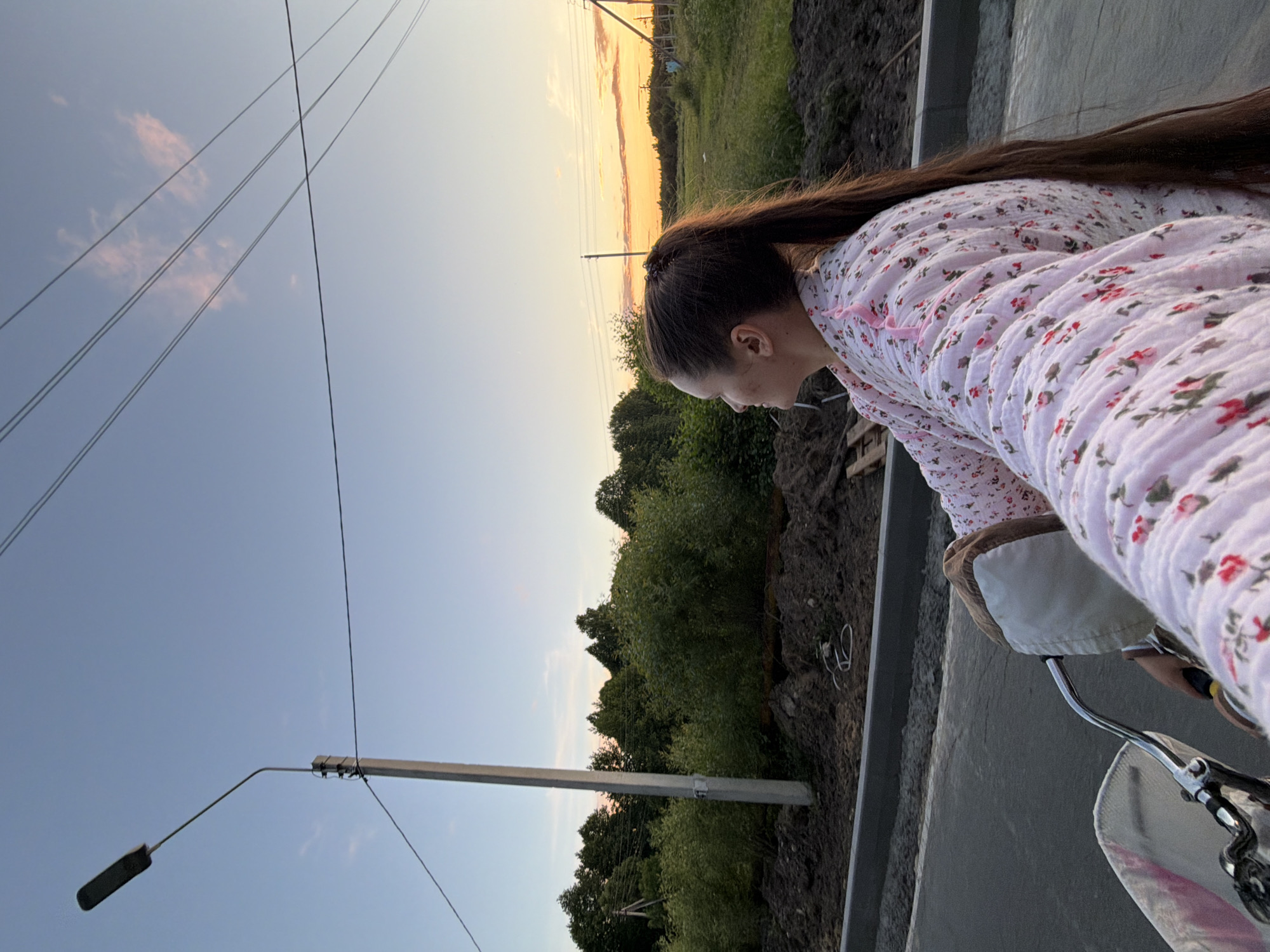A woman with long dark hair tied in a ponytail rides a bicycle on a paved road, looking to her right. She wears a light pink robe with a floral pattern. The road is under construction, with piles of dirt and a concrete curb visible. In the background, there is a line of trees and fields. Power lines and a street lamp are also present. The sky is clear with a few wispy clouds, and the sun is setting, casting a warm orange glow on the horizon. The location is the city of Krasnyy Mayak, Russia.