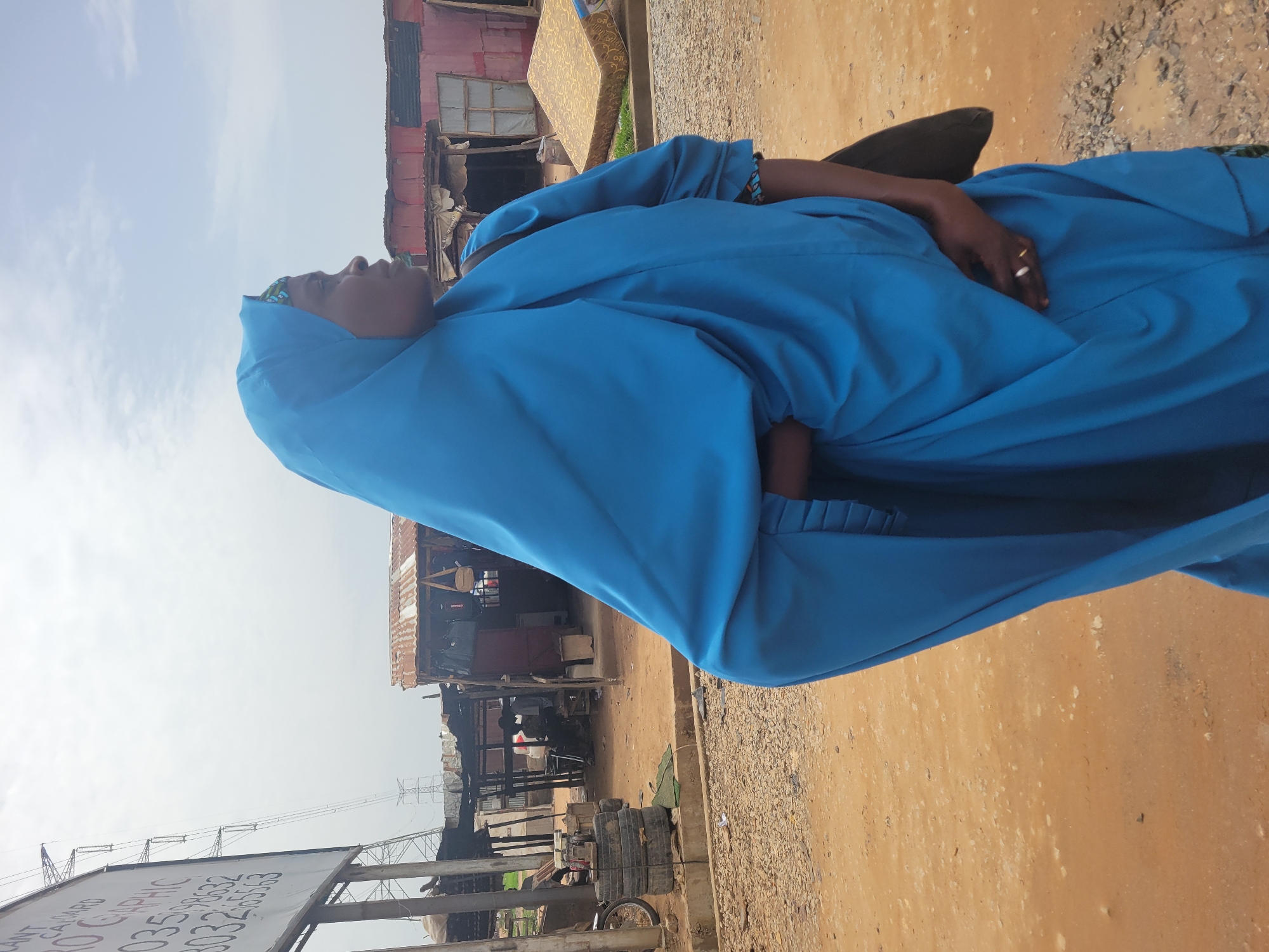 A woman in a blue garment stands on a dirt road in Kwamba, Nigeria. Background: shacks and utility poles.