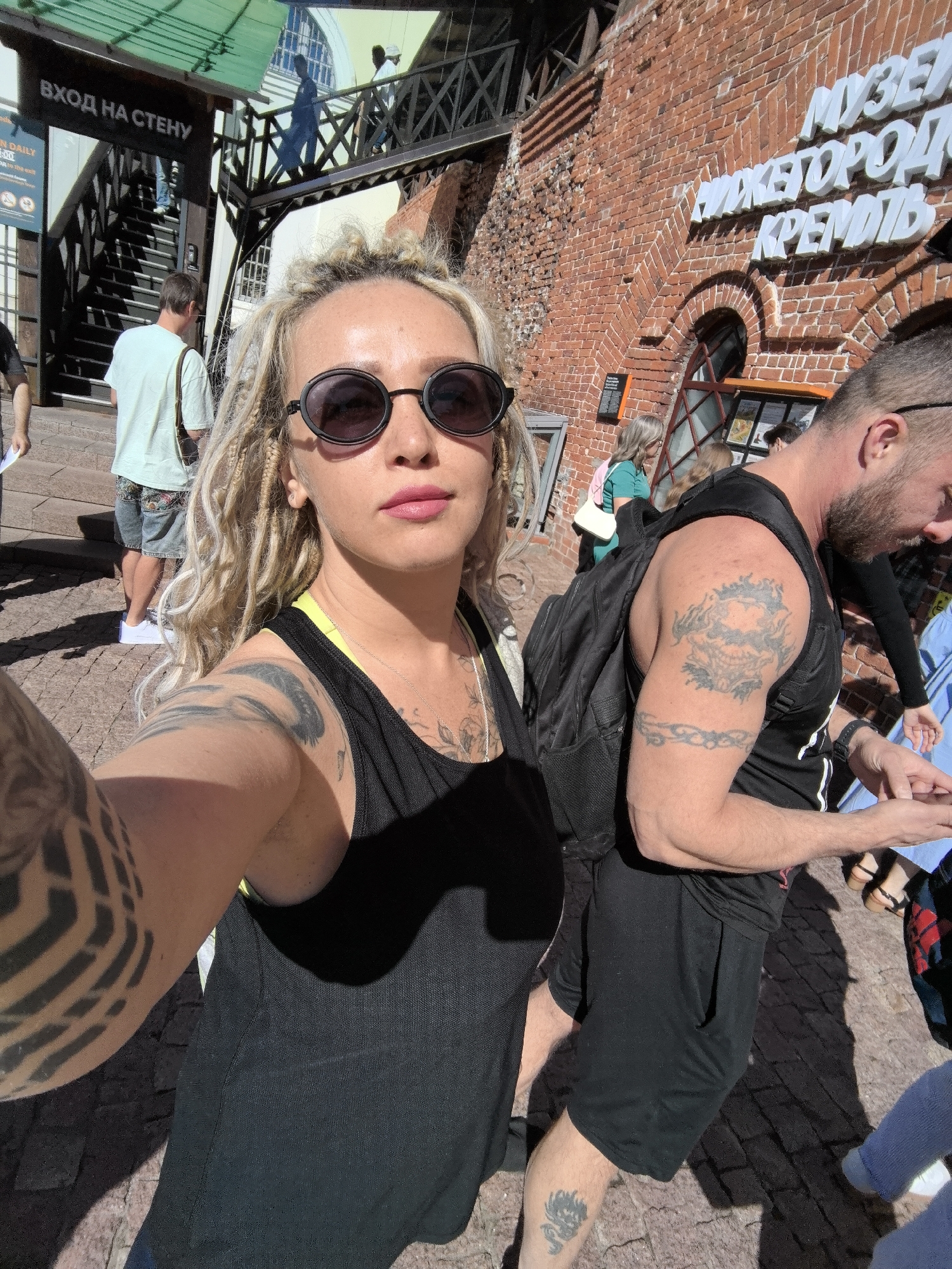 The image captures a selfie taken by a woman with blonde dreadlocks and extensive arm tattoos, wearing a black tank top and sunglasses. She is positioned in the foreground, with a brick building featuring signage in Russian visible behind her. The signage appears to be for a museum or historical site, with the words "МУЗЕЙ" (Museum) and "НИЖЕГОРОДСКИЙ КРЕМЛЬ" (Nizhny Novgorod Kremlin) discernible. To the left of the woman, a stairway leads upwards with a sign reading "ВХОД НА СТЕНУ" (Entrance to the Wall). Several other people are present in the background, including a man with tattoos on his arm and a backpack, and a man in a light blue t-shirt. The setting appears to be an outdoor historical or cultural attraction in Nizhny Novgorod, Russia, on a sunny day, judging by the shadows. The ground is paved with what looks like cobblestones or similar material.