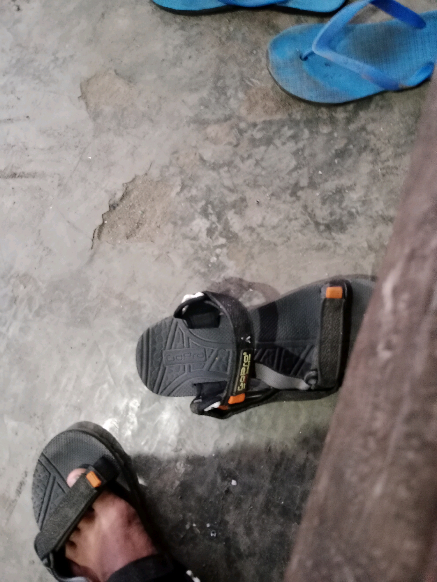 In Ringinrejo, Indonesia, a pair of dark gray "GoPro" branded sandals are visible on a concrete floor. One sandal is worn on a person's left foot, with the toes peeking out. The other sandal is positioned beside it. In the background, a pair of bright blue flip-flops are also present on the same concrete surface. The floor appears worn and slightly dirty.