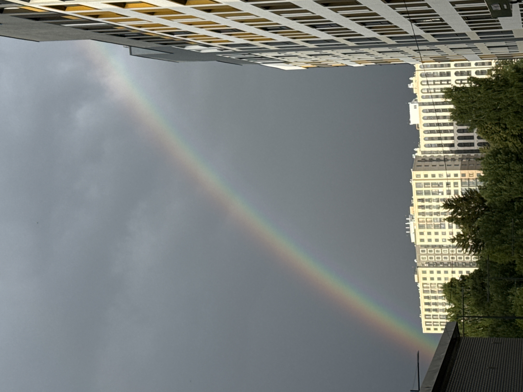 This media file captures a striking urban scene featuring a rainbow against a gray, overcast sky. Two tall, modern apartment buildings dominate the frame, one on the left and another on the right, creating a sense of depth and urban density. The building on the left is predominantly gray with a facade of white and yellow window frames, arranged in a repeating grid pattern. The building on the right is lighter in color, with a similar grid of windows, and is partially obscured by lush green trees.
A vibrant rainbow arcs across the sky, its colors clearly visible against the somber backdrop of clouds. The rainbow appears to be a primary arc, with its spectrum of colors transitioning from red at the top to violet at the bottom, as it stretches from the upper left of the frame towards the lower right.
The perspective suggests the photo was taken from a low vantage point, looking upwards between the buildings. The orientation of the image is rotated, making the buildings appear to lean inwards from the top and bottom. The presence of the rainbow indicates that it is likely raining or has recently rained, and the sun is out, suggesting a transient weather condition typical of a passing shower. No people are visible in the image, and no text is present. The scene evokes a sense of natural beauty emerging from an urban environment, perhaps conveying a feeling of hope or wonder. The location context of Izhevsk, Russia, provides a geographical anchor to this atmospheric display.