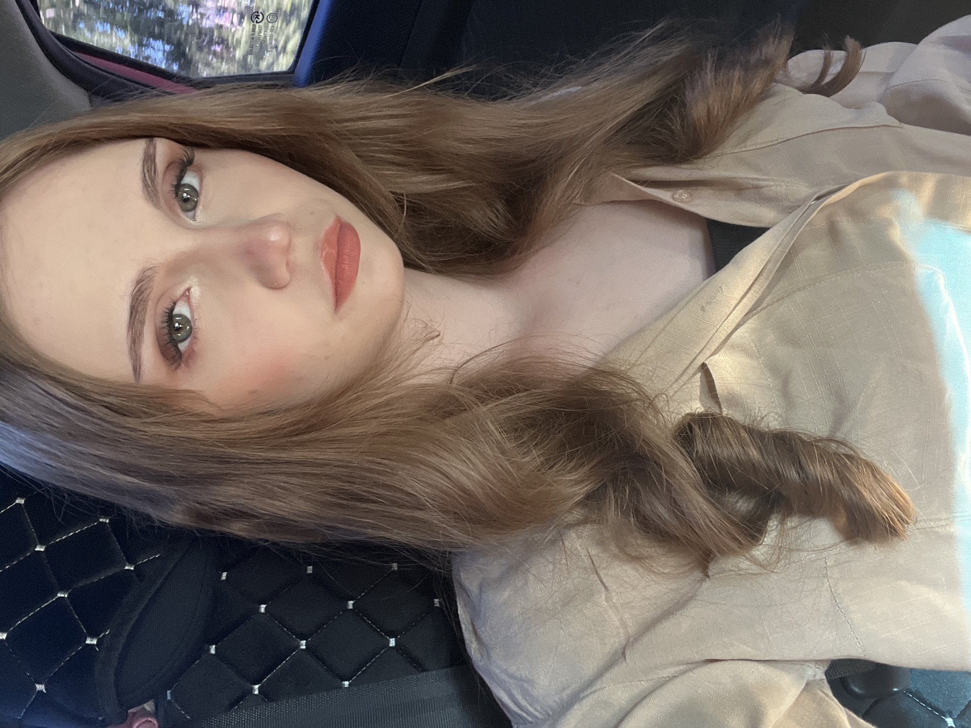A young woman with long, wavy, light brown hair is shown from the chest up, looking forward. She is seated in the passenger seat of a vehicle, as indicated by the car seat behind her and the window with a blurred outdoor scene to her right. She is wearing a light beige button-up shirt. Her makeup includes eyeshadow and lipstick. The scene appears to be captured during daylight hours. There is no discernible evidence to suggest this image was taken in the city of Новая Отрадовка, Russia.