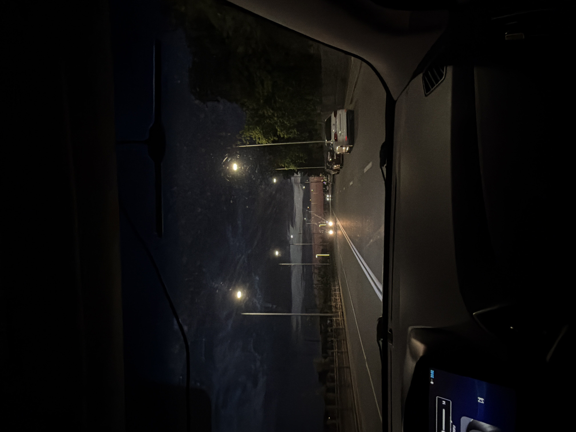 The image shows a nighttime view from the interior of a car traveling on a road in Ufa, Russia. The sky is dark with clouds, illuminated by streetlights and the headlights of other vehicles. The road is paved and has double white lines marking the lanes. Several cars are visible on the road, either parked or moving. Trees and other vegetation line the right side of the road. The dashboard of the car is partially visible, showing a display screen. The scene appears to be a typical urban or suburban environment at night.