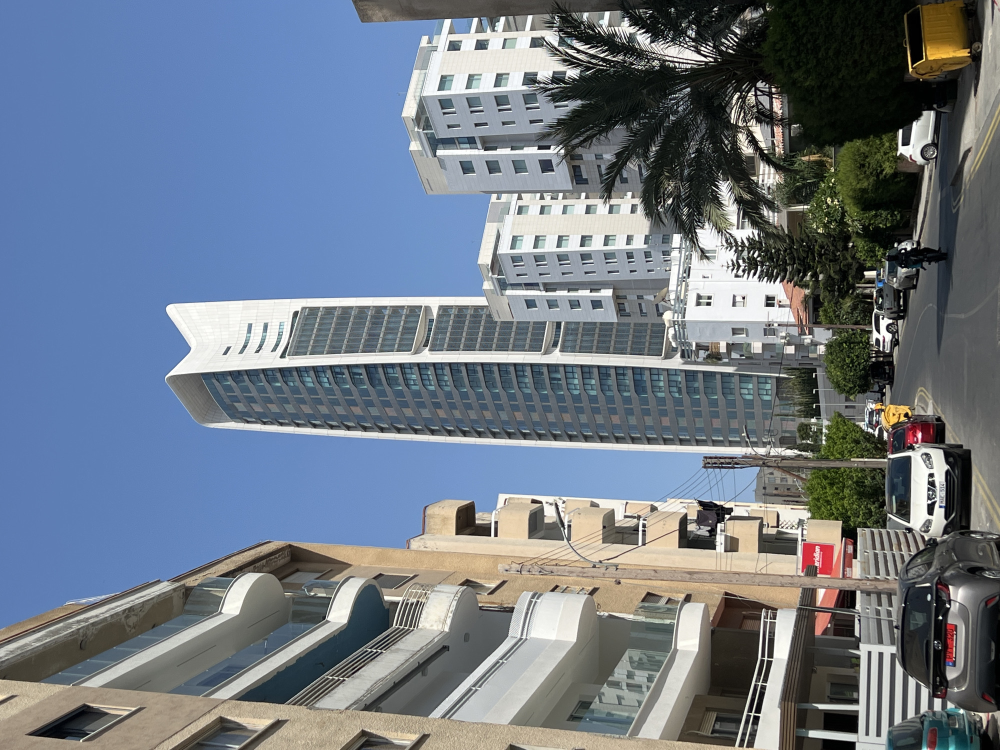 This is a vertical, eye-level, outdoor photograph taken on a sunny day. The scene is a street with several modern buildings and parked cars. In the foreground, a multi-story building with a beige facade and white balconies is visible. Behind it, a very tall, sleek skyscraper with a curved, white top and numerous reflective windows dominates the background. To the right of the skyscraper, another building with a white facade and regularly spaced windows is seen. Further down the street, on the right side of the frame, several cars are parked, including a white SUV, a gray SUV, and a yellow dumpster. A motorcycle with a rider is also present on the street. Trees and other greenery are interspersed between the buildings. The sky is a clear, bright blue, suggesting pleasant weather. The image appears to have been taken in Limassol, Cyprus, given the location context. No specific activity or event is clearly depicted, other than the general street scene. One piece of visible text reads "Meridian" in red on a white sign.
