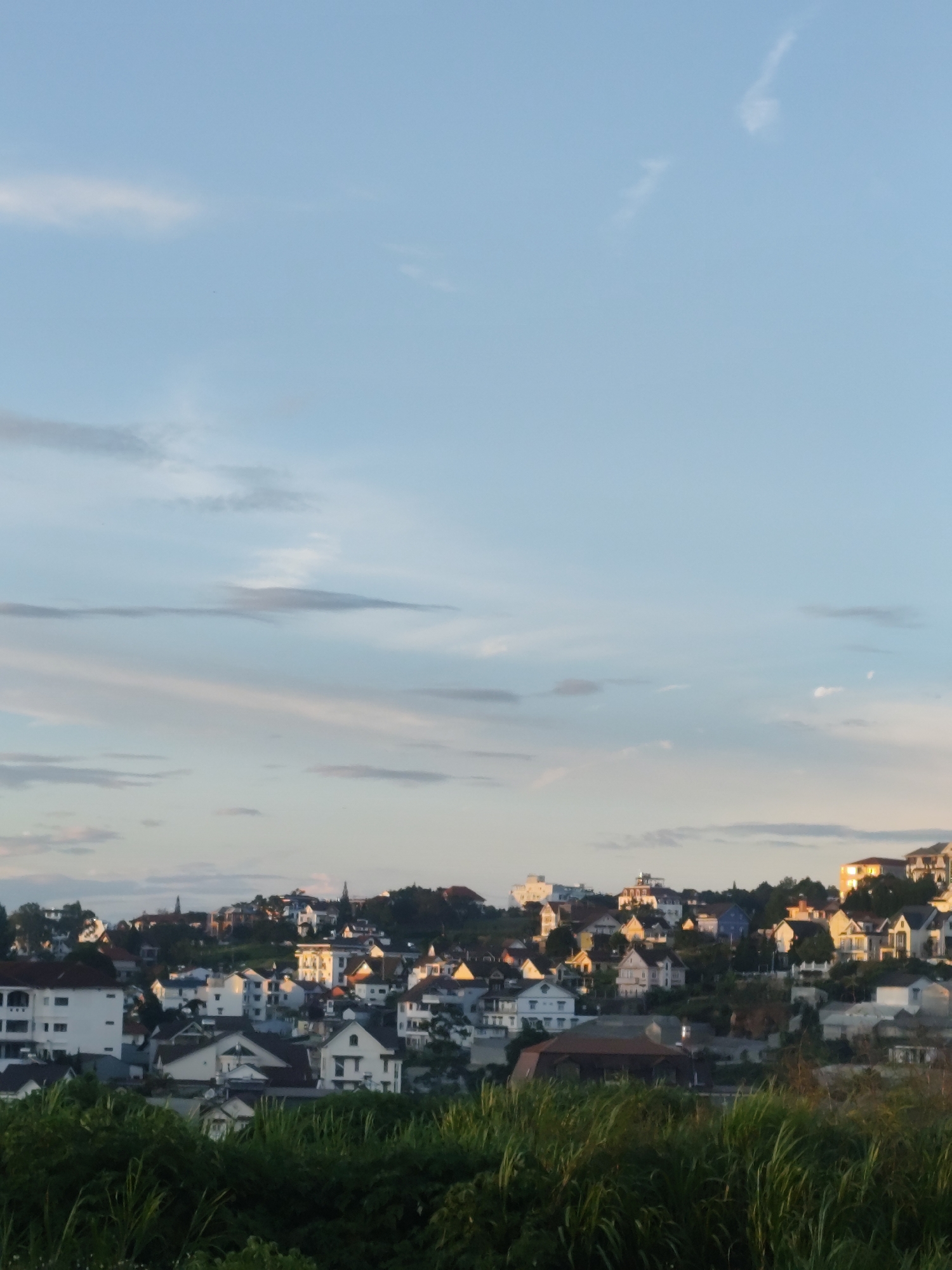 This media file depicts a panoramic view of a hilly town, likely Da Lat, Vietnam, at sunset. The foreground is dominated by lush green grass, suggesting a natural vantage point overlooking the settlement. The town itself is a collection of houses and buildings clustered on rolling hills, with varying architectural styles and roof colors. Some houses are brightly colored, while others are white, contributing to a picturesque and somewhat whimsical scene. The setting sun casts a warm, golden glow on the buildings, particularly on the upper right portion of the image, highlighting the contours of the landscape.
The sky above is a soft gradient of blue and pale orange, with wispy clouds scattered across it, indicative of a calm and pleasant evening. The overall atmosphere is serene and tranquil, evoking a sense of peace and quietude. The time of day is clearly sunset due to the warm lighting and the colors of the sky. While there are no visible people or specific objects in the foreground, the focus is on the interplay of light and shadow on the urban landscape and the natural beauty of the surroundings. The scene suggests a residential area with a blend of traditional and modern housing styles, nestled amidst the natural topography of the hills. The presence of abundant greenery in the foreground further emphasizes the natural setting of the town. No text is visible in the image.