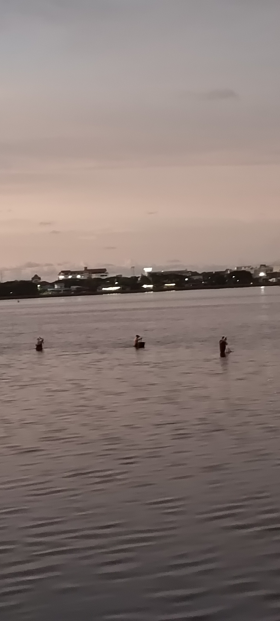This image depicts a serene dusk scene over a body of water, with a distant city skyline providing a backdrop. The sky is a soft gradient of gray and pale orange, suggesting the twilight hours. The water is calm, with gentle ripples reflecting the muted light.
In the water, three individuals are partially submerged, appearing to be engaged in some activity. The first person on the left stands in the water, possibly holding something. The second person, closer to the center, is also standing and seems to be interacting with a dark, rectangular object. The third person on the right is wading in the water, holding what looks like a net or a fishing implement. Their postures suggest they are either fishing or collecting something from the water.
The distant shoreline is lined with buildings, some of which are illuminated, indicating it is a populated area. The overall impression is one of quiet activity during a peaceful transition from day to night. The location is Lhokseumawe, Indonesia. There is no visible text in the image.