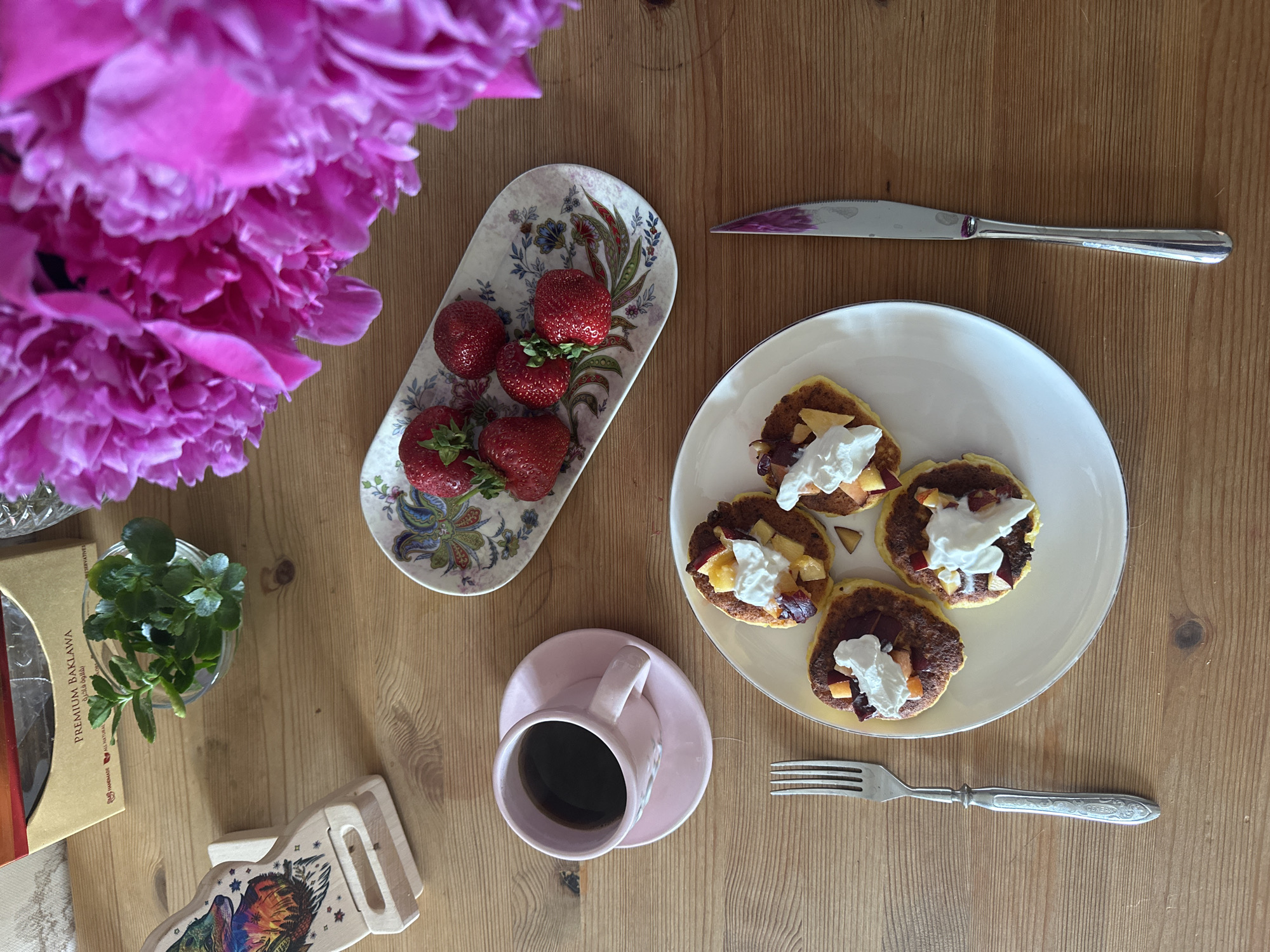 This is a top-down, medium shot of a breakfast spread on a wooden table. In the upper left is a box of "PREMIUM BAKLAWA". To the right of the box is a small glass bowl with green herbs. To the right of the herbs are vibrant pink peonies. Below the baklava box is a wooden phone stand with a colorful fox design. In the center left, a pink teacup and saucer holds black coffee. To the right of the teacup is a small oval plate with several strawberries. In the center right, a white plate holds four small pancakes topped with whipped cream and fruit. A silver fork rests to the left of the white plate, and a silver knife with a purple stained blade rests to the right. The table surface is made of light brown wood planks. No people are visible in the image. This scene is not specifically located in Vladimir, Russia.