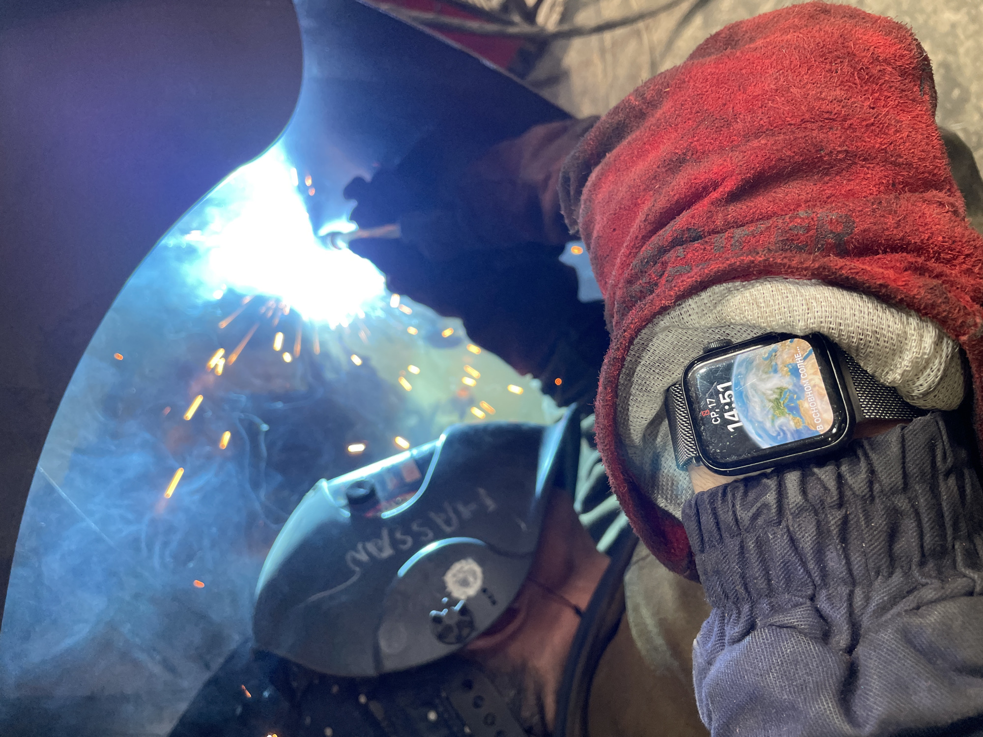 The image depicts a person welding in what appears to be an industrial or workshop setting. The primary subject is a welder wearing protective gear, including a welding mask with the name "HASSAN" written on it in white marker, and heavy-duty gloves. One hand, covered by a red and grey glove, is positioned near the welding torch, emitting a bright, intense light and sparks. The other hand, also gloved, is visible in the foreground, wearing a smartwatch. The watch face displays a globe image, the time "14:51," the date "СР. 17" (likely Wednesday, 17th), and some Cyrillic text which reads "ВОСНОВНОМ СОТЕ". Blue smoke or fumes from the welding process are also visible. The lighting is dominated by the bright glare of the welding arc, creating a high-contrast scene with deep shadows. The overall impression is one of focused, industrial work. The location context provided is Tomba, Montenegro.