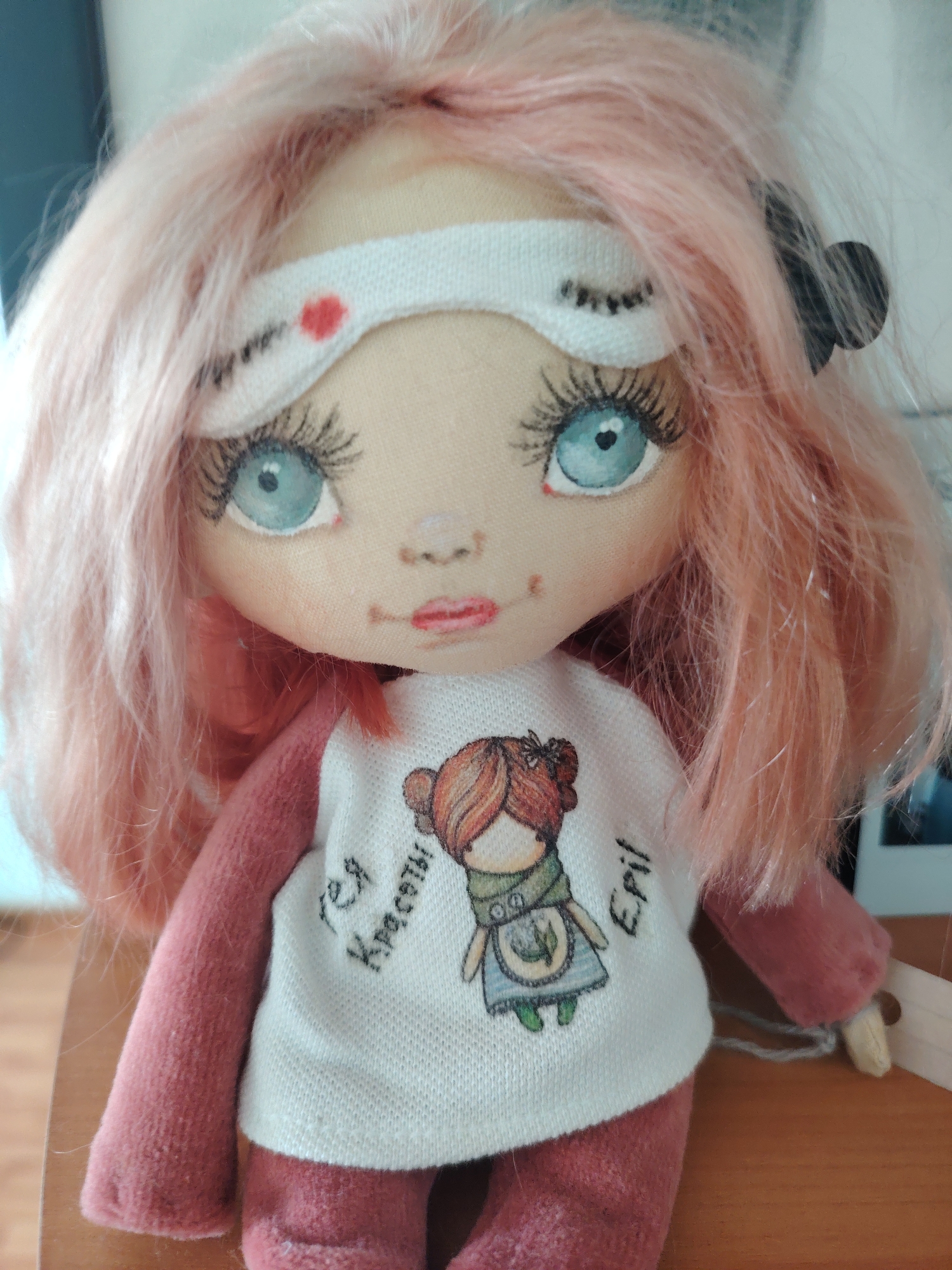 A close-up, eye-level indoor shot features a handmade cloth doll with long, wavy, pastel pink hair. The doll wears a white sleep mask with black painted eyelashes and a small red dot on the forehead. Its eyes are large, bright blue with black pupils and long, dark eyelashes. The doll has a small, pink, smiling mouth and lightly blushed cheeks. It is dressed in a white long-sleeved shirt with a drawing of a small figure in an apron and a green scarf, and the words "Tea Krasoty" and "Epil" are embroidered on the shirt. The doll's lower body is dressed in pink. The doll is positioned on a wooden surface, possibly a table or shelf. The background is blurred, showing hints of indoor elements and light. There is no specific information to determine its location in Konakovo, Russia.