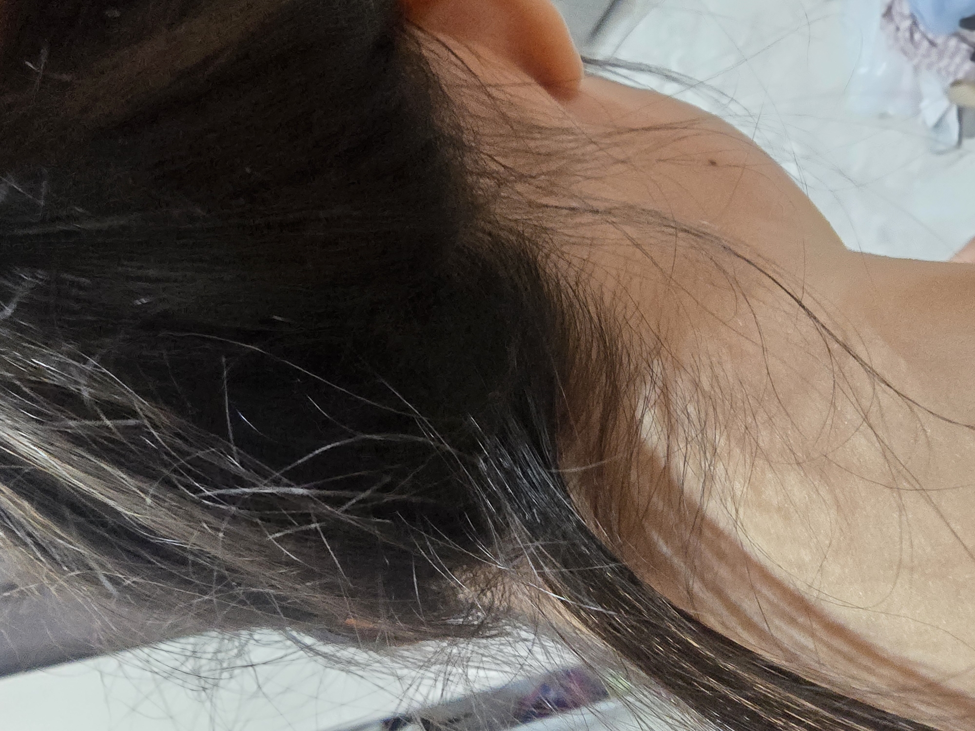 The image shows a close-up of a person's dark hair and neck. The lighting suggests an indoor environment, possibly a home, given the presence of what appears to be a patterned textile in the background. The focus is on the texture of the hair, with individual strands visible, some appearing lighter or thinner than others. The skin of the neck is also visible, showing some texture and a small dark mole. The overall impression is a candid, personal shot, not a public or identifiable urban scene. There is no information in the image that can be used to determine the location in Ulan-Ude, Russia.