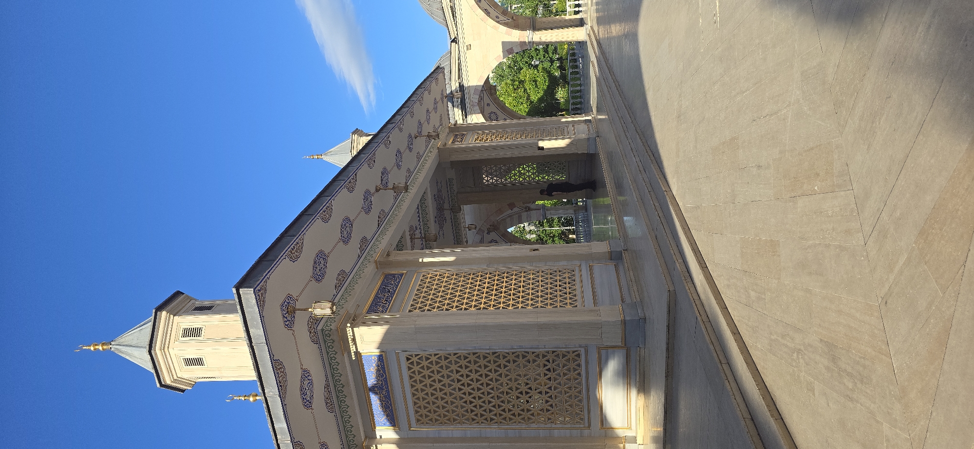 The image shows an ornate arcade with Islamic architectural details, likely part of the Heart of Chechnya Mosque complex in Grozny, Russia. The structure features decorated ceilings, archways, and latticework screens. Two individuals are present within the arcade. A clear, blue sky is overhead. The foreground consists of a stone-paved walkway.