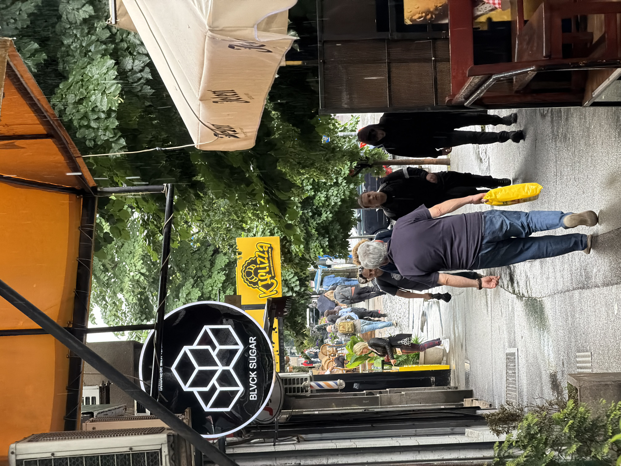 The image depicts a street scene in Belgrade, Serbia. The foreground features signs for "BLVCK SUGAR" and "KIPizza" hanging over the sidewalk. An air conditioning unit is visible on the building. A group of people is walking along the narrow street, including a man in jeans carrying a yellow bag. A barber pole is also visible among the storefronts. The street is wet. Trees line the street, providing partial shade. Awnings and canopies extend from the buildings over the sidewalk. Chairs and tables appear to be set up outside a restaurant.