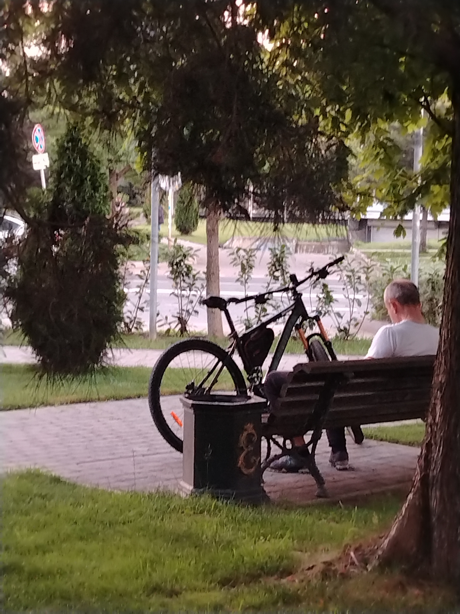 A man is sitting on a park bench, facing away from the camera. He is wearing a white t-shirt and dark pants. Next to him is a bicycle. The scene is set in a park with trees and a paved walkway. In the background, there is a road and some greenery. The image appears to have been taken during the daytime. The provided metadata indicates that the location is Krasnodar, Russia.