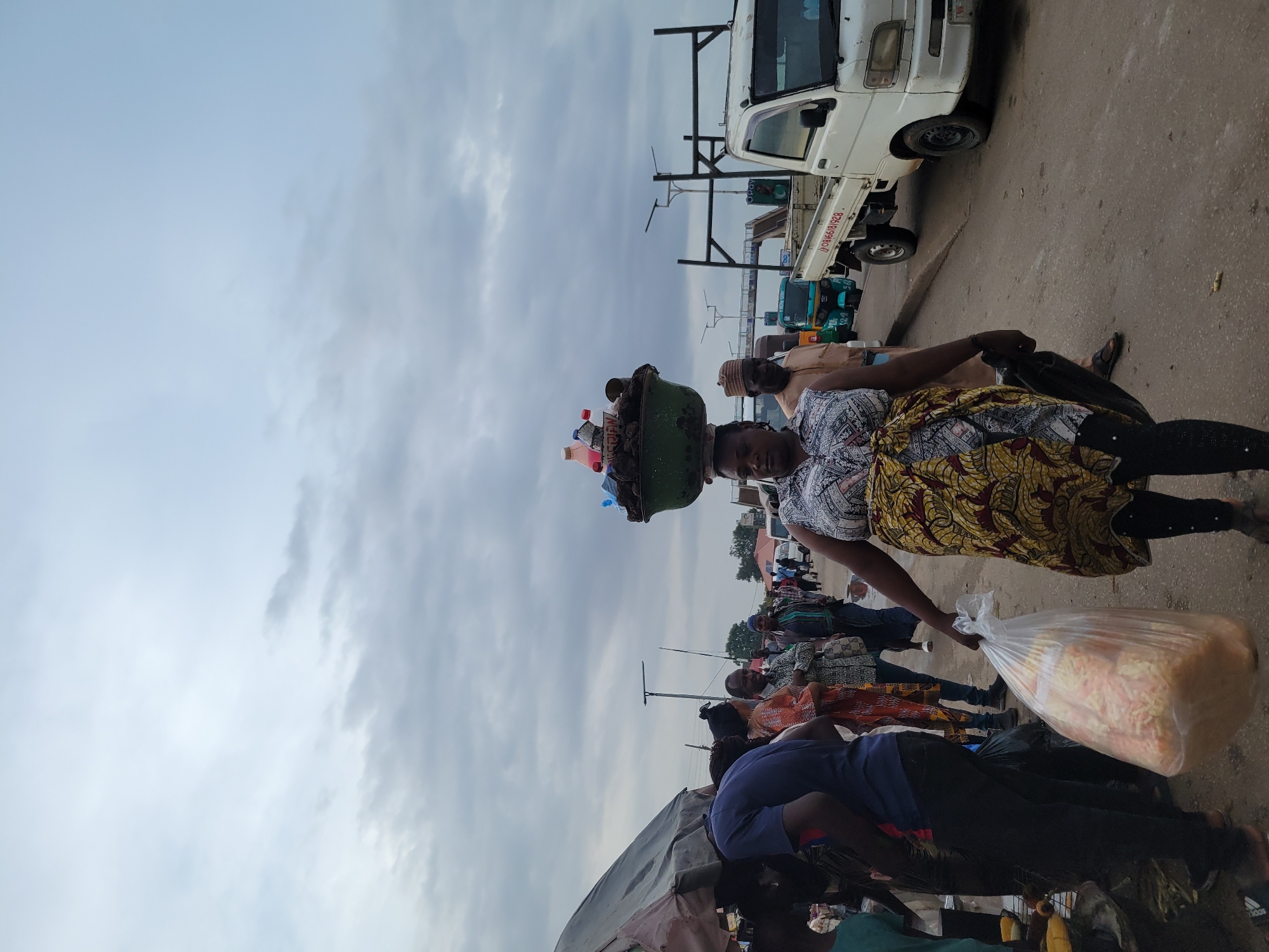 The image depicts a crowded street scene in Gura Top, Nigeria. A woman carries a large container on her head, filled with various items including what appear to be plastic containers and packaged goods. She is holding a large plastic bag filled with food items in her hand. Behind her, other people are visible, some standing and some appearing to be seated or kneeling by what might be market stalls. There are vehicles parked on the side of the street, including a white truck. The sky is overcast.
