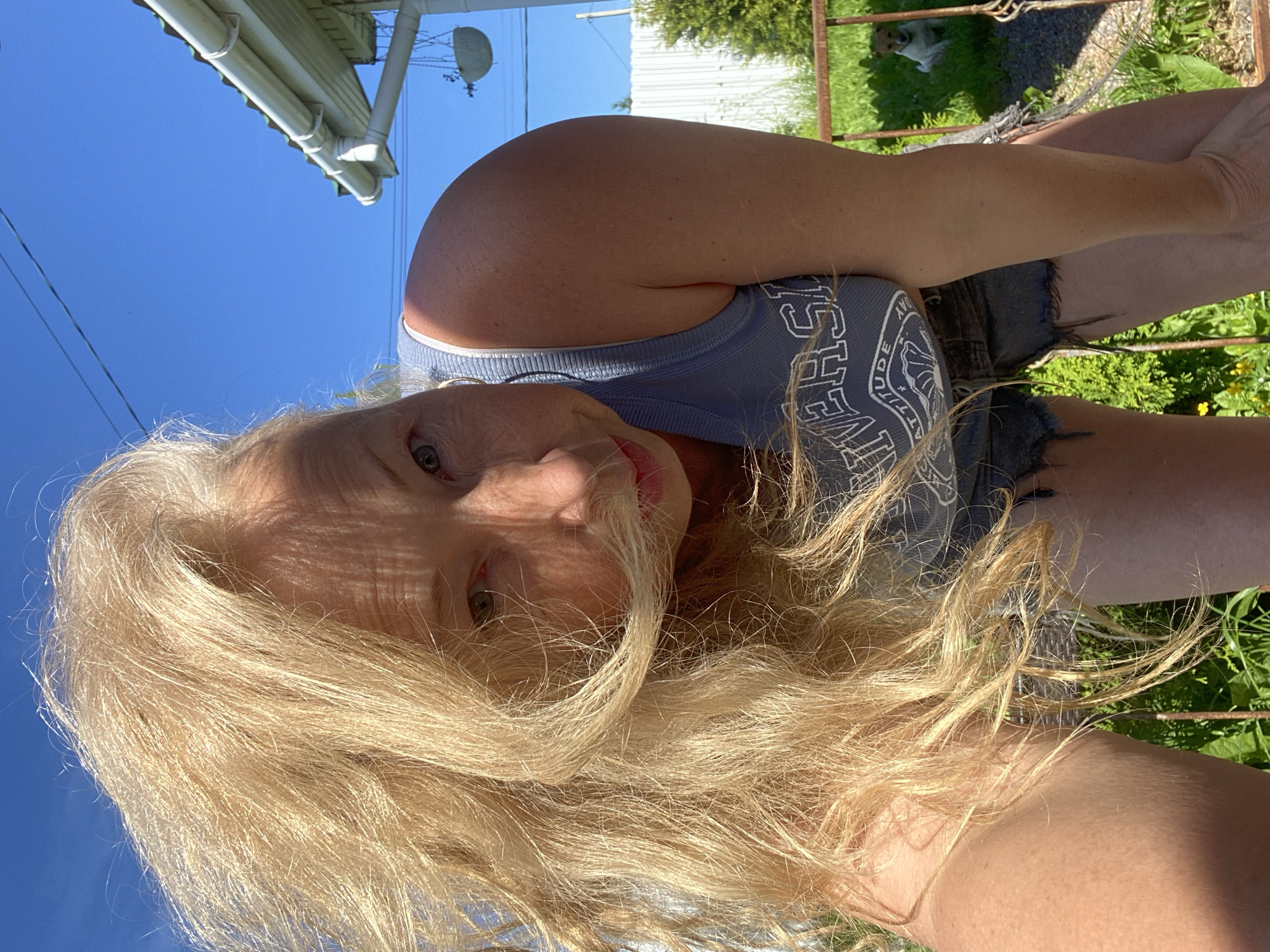The image shows a woman with long blonde hair, looking at the camera. She is wearing a blue tank top and denim shorts. The photo is taken outdoors, with a clear blue sky in the background. There is a white building with a green roof and gutters on the left side of the frame. A metal fence is visible on the right, with green foliage behind it. A small dog is sitting behind the fence. The lighting suggests it is daytime, likely sunny. The overall mood appears casual and relaxed. Visible text on the woman's tank top includes "HUDE" and "DAY". The location context provided is Bol'shoe Zaborod'e, Russia.