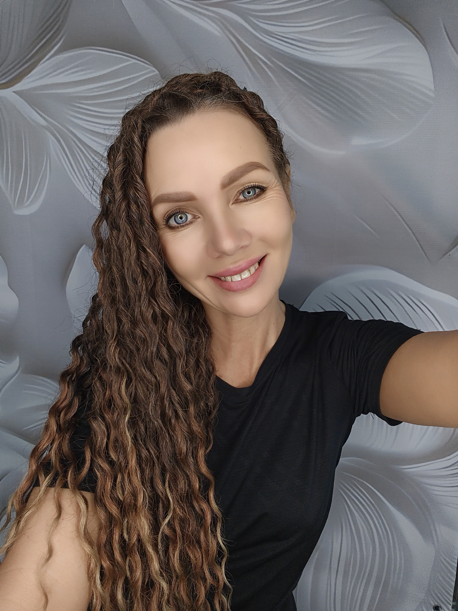 A medium shot selfie shows a woman with long, curly brown hair and blue eyes smiling. She is wearing a black t-shirt and stands in front of a grey background with a floral pattern.