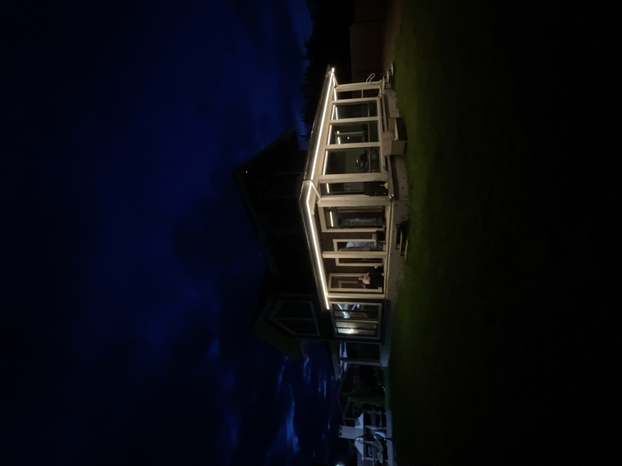 An exterior shot of a building at night, potentially located in Solov'evka, Russia. The building has a glass enclosed portion with illuminated white lights outlining its structure. The left of the frame features a dark blue sky, while the right shows dark green lawn. In the reflection of the windows, a person can be vaguely seen. Below the building, there are dark shapes which may be furniture or yard decorations.