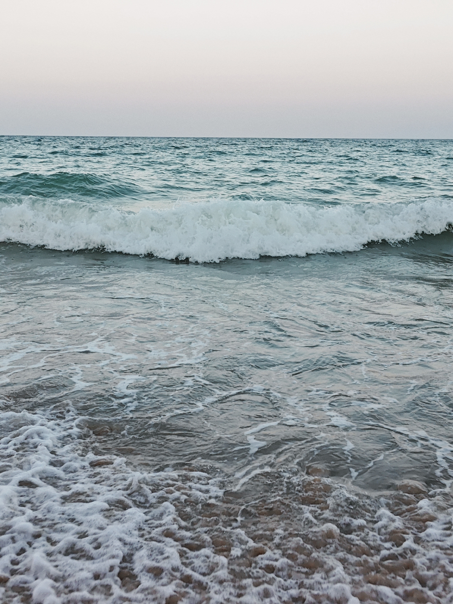 The image displays a tranquil seascape with a gentle wave breaking on a sandy shore. The sky is a soft, pale pinkish-gray, suggesting either dawn or dusk. The ocean water is a cool turquoise color, with ripples and small whitecaps visible. The wave in the foreground is frothy and white as it approaches the shore, which is composed of small pebbles and sand, with patches of foam from the receding water. The scene is devoid of any people or man-made objects, creating a natural and serene atmosphere.
