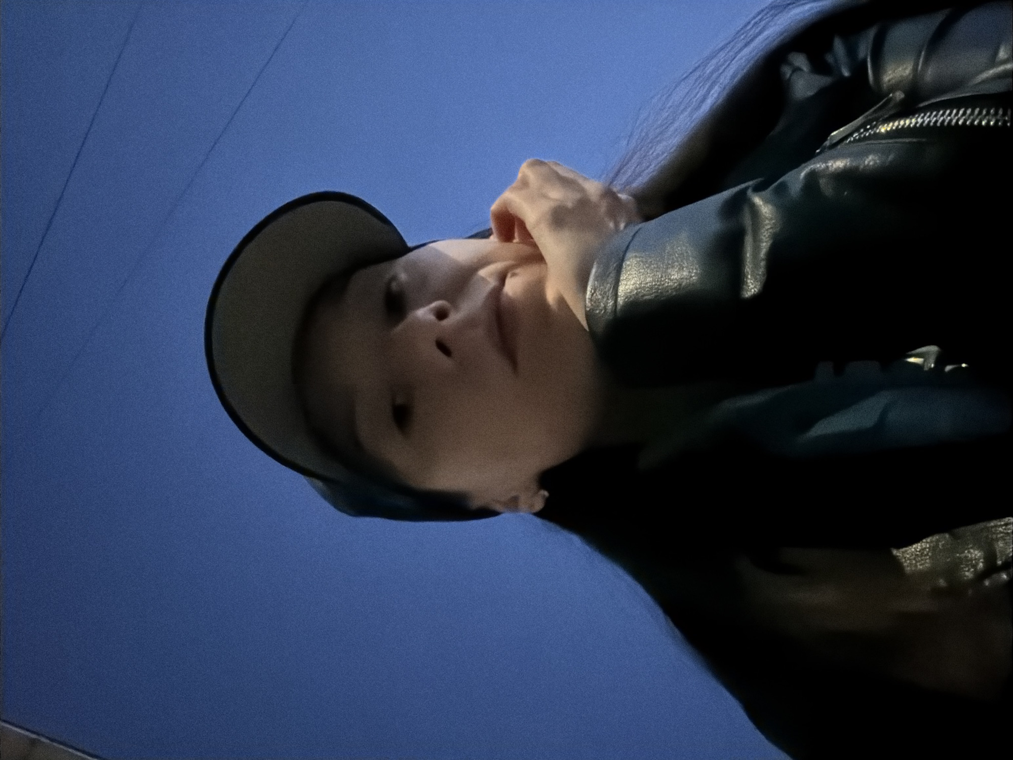 A low-angle, slightly blurry selfie shows a young woman wearing a black baseball cap and a dark, possibly leather, jacket. Her left hand is supporting her chin, and her right hand is visible near her ear. The background is a deep blue sky, suggesting it is dusk or nighttime. There are a few faint, parallel lines in the sky, which could be power lines. The image does not contain any specific landmarks or details that would allow for identification of the location within Raevka, Russia.
