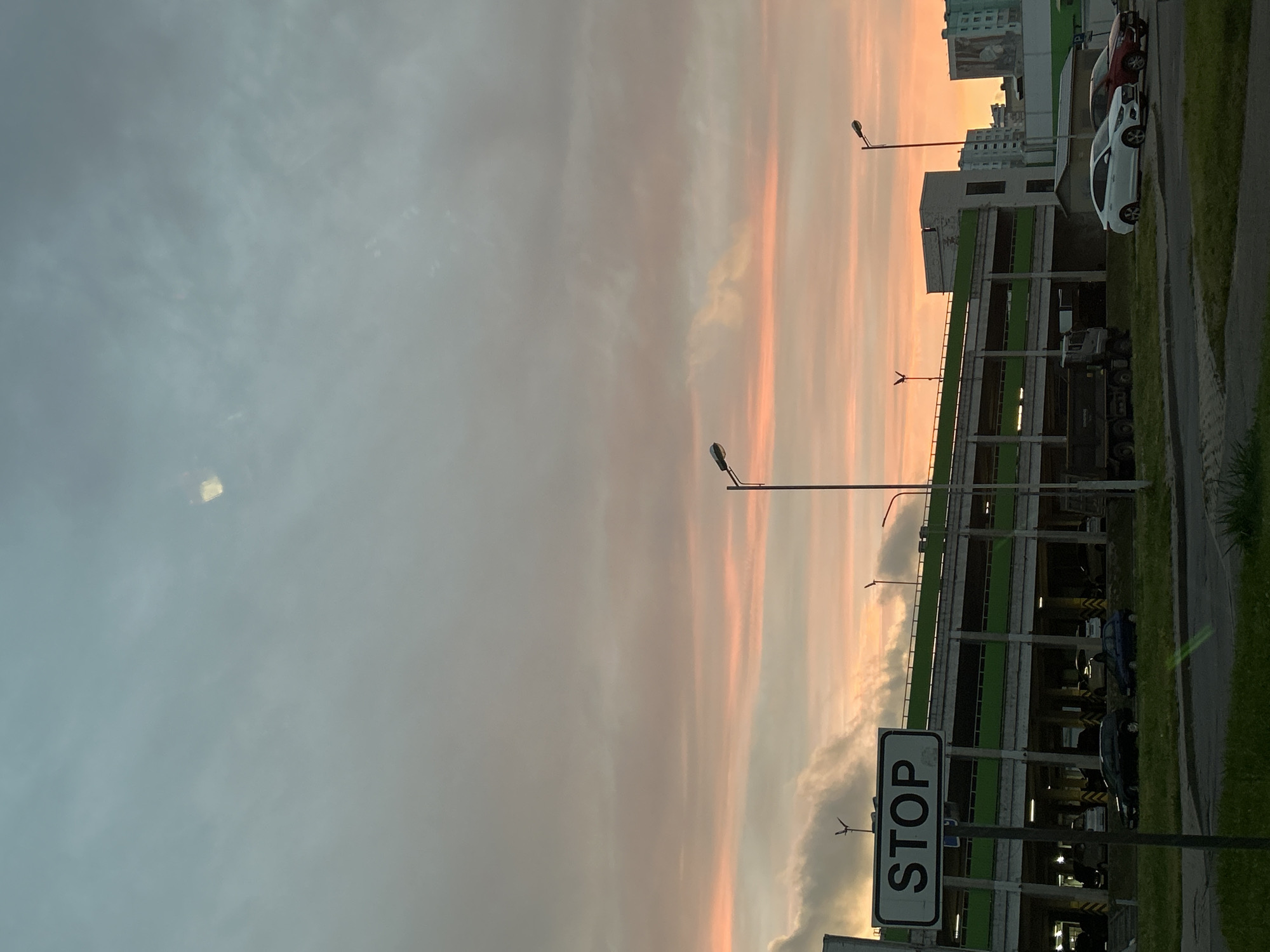 The image captures an outdoor scene at dusk in Minsk, Belarus. A multi-level parking garage with green accents is prominent in the mid-ground. In the foreground, a stop sign on a pole is clearly visible on the left side. Several parked cars are scattered throughout the scene, including a white sedan and a red sedan on the right. A dark-colored truck is positioned in front of the parking garage. The sky is filled with dramatic clouds, tinged with shades of orange and pink, indicating a sunset. The overall environment appears to be an urban setting, likely a residential or commercial area.