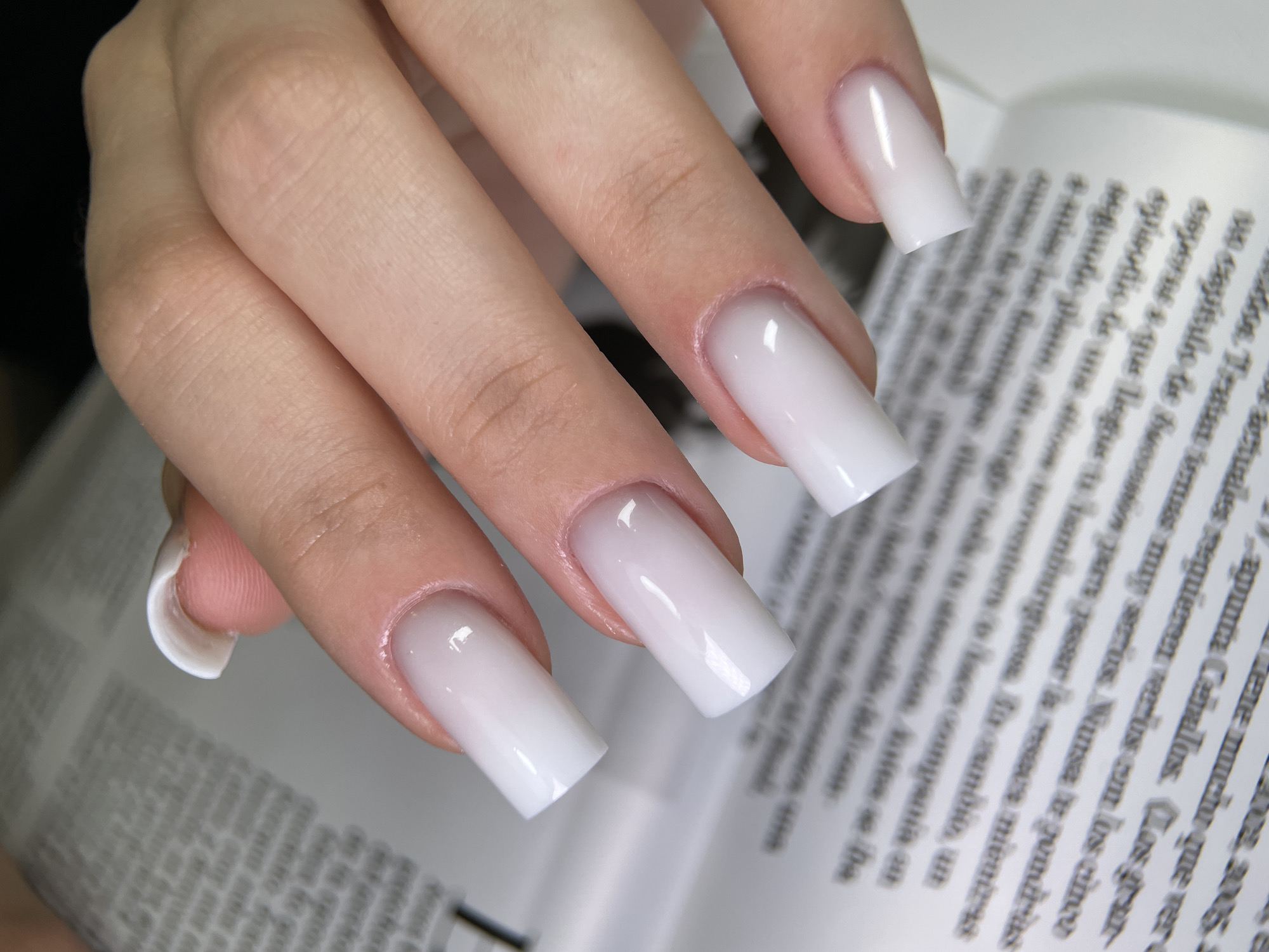A close-up shot, taken in Izhevsk, Russia, features a hand with light-toned skin resting on an open book or magazine. The subject's fingers are adorned with long, square-shaped nails showcasing a milky white ombré or "milky bath" manicure, transitioning from a translucent, natural hue near the cuticles to an opaque white at the tips. The nails appear glossy and well-maintained. The background, largely out of focus, reveals pages of a book or magazine filled with text. The overall lighting is soft and even, highlighting the details of the manicure.
Visible text on the open pages includes the year "2005" and several words in a Romance language, likely Spanish or Portuguese, such as "capítulo," "mientras," "teatral," "nunca," "hamburguesa," "episodio," "compañía," "actuales," "versus," "apunta Ceballos," and "mucho que ver." Some parts of the OCR text appear garbled, but the overall context suggests articles or stories are printed on the pages. The activity depicted is primarily a display of the manicure, possibly while engaging with or posing with reading material.