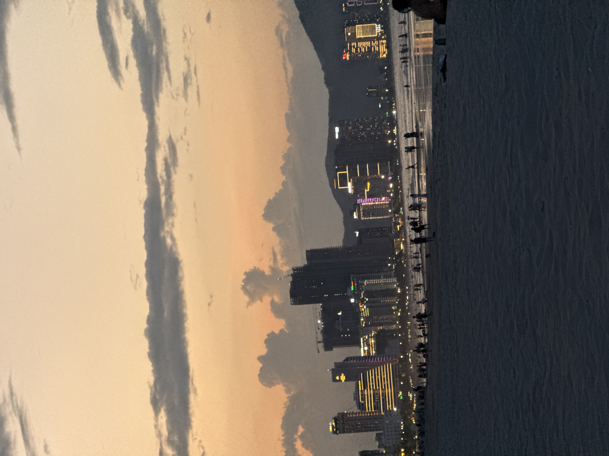 This image presents a panoramic view of a coastal city, identified as Đà Nẵng, Vietnam, at dusk or early evening. The sky, occupying the upper portion of the frame, transitions from soft orange and yellow hues near the horizon to lighter shades, interspersed with long, streaky dark gray clouds.
Below the expansive sky, the city's skyline unfolds, showcasing numerous modern high-rise buildings. Many of these structures are brightly lit with various artificial lights, casting a glow and highlighting their architectural forms against the dimming light. Some buildings display distinctive patterns of light, suggesting commercial or residential use. A dark, undulating silhouette of mountains or hills can be seen in the background, behind the urban sprawl.
In the foreground, a wide sandy beach or promenade stretches horizontally. The silhouettes of many people are visible walking, standing, and strolling along this area, enjoying the evening by the water. To the right of the beach, a vast body of calm, dark water, likely the ocean, extends into the distance. The overall atmosphere is serene and leisurely, characteristic of an evening spent by the coast in a vibrant city. There is no discernible text visible in the image.