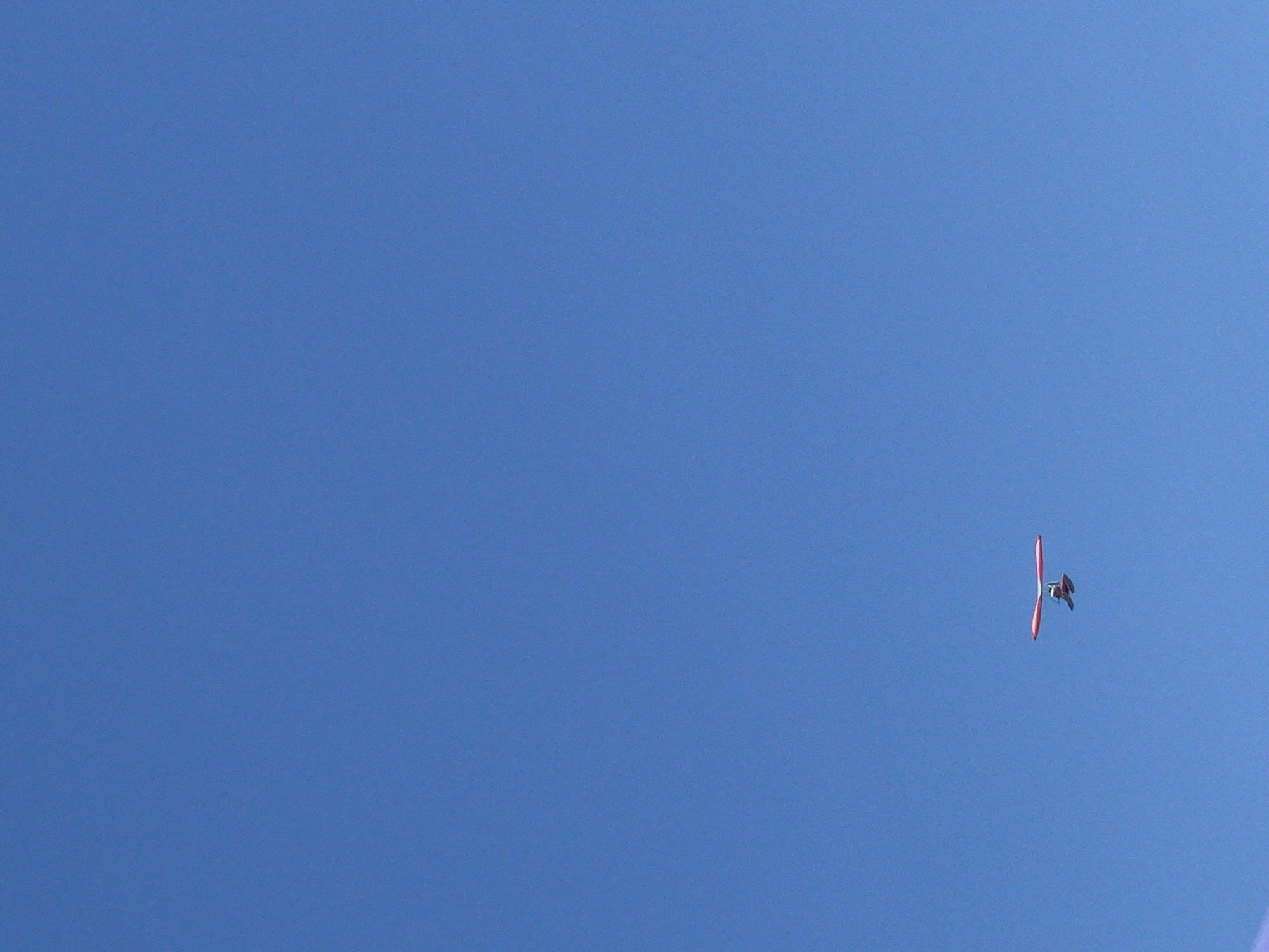 A hang glider is shown in flight against a clear blue sky. The hang glider has a red wing and a dark body. It is a distant object and its details are not clearly discernible. The scene is set in the open sky above Bosteri, Kyrgyzstan. There are no other objects or people visible in the frame.