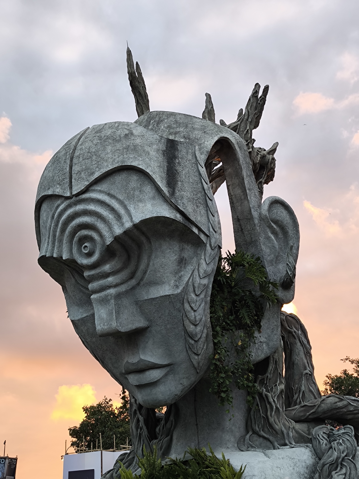 The image shows a close-up of a large, gray, stone sculpture of a stylized human head against a sky with pink and orange hues, indicative of sunset. The sculpture features intricate details, including a spiraling pattern on the forehead, a large ear, and vine-like structures integrated into its form. Lush green plants are growing on and around the sculpture, suggesting it is outdoors and has been there for some time. In the background, silhouetted trees and some construction scaffolding are visible. The sculpture is located in Kediri, Indonesia, specifically at the GUPALA sculpture park. The overall impression is one of an artistic installation blending natural and human-made elements.