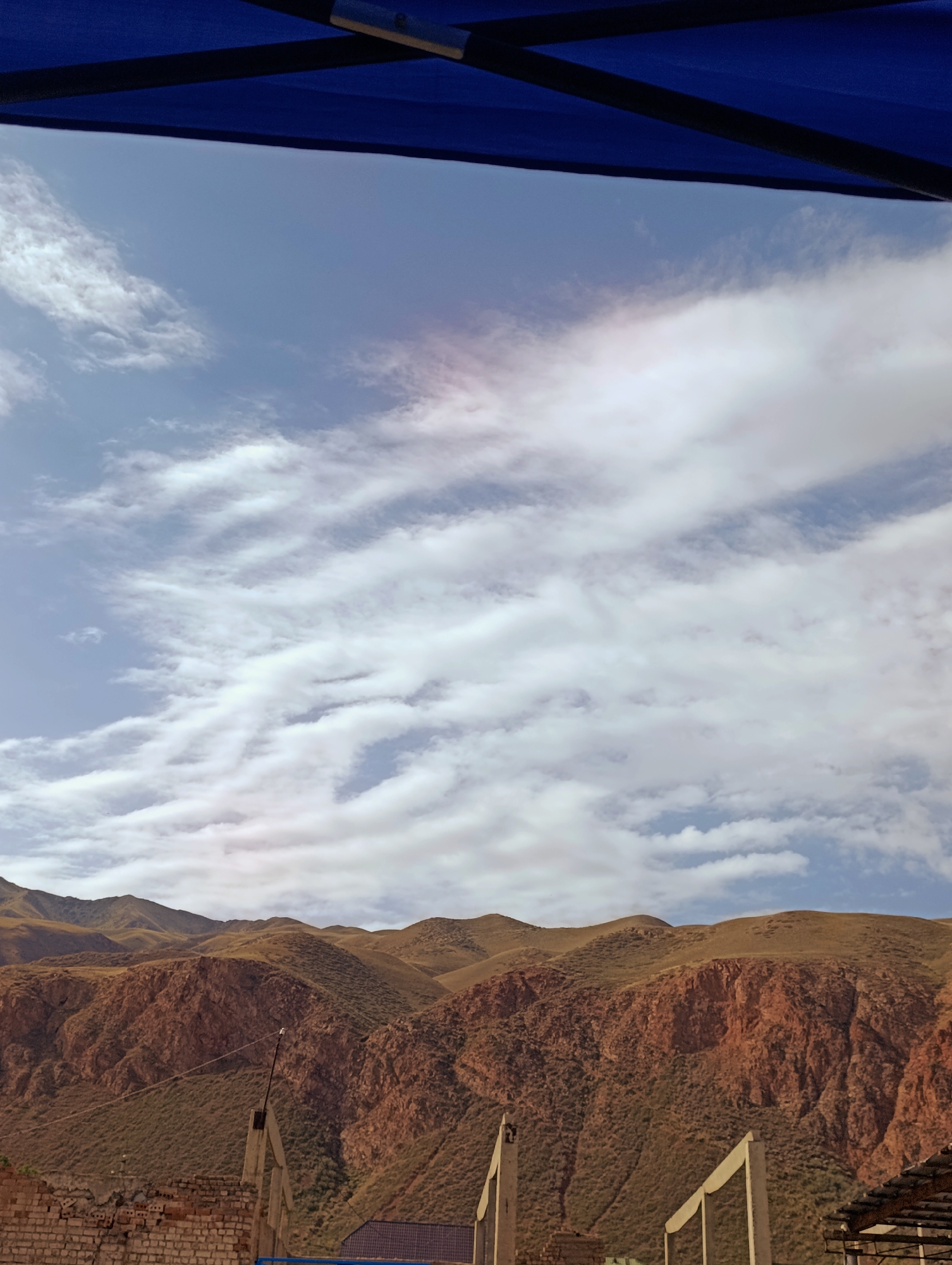 An outdoor scene in Naryn City, Kyrgyzstan, features a view of rugged, brown-and-red toned mountains under a partly cloudy sky. The sky is a pale blue, streaked with wispy white clouds. In the foreground, a dark blue canopy is visible, casting a shadow. Below the mountains, there are hints of vegetation and some man-made structures, including a brick wall, concrete pillars, and the edge of a roof. The overall impression is of a natural landscape with some human presence.