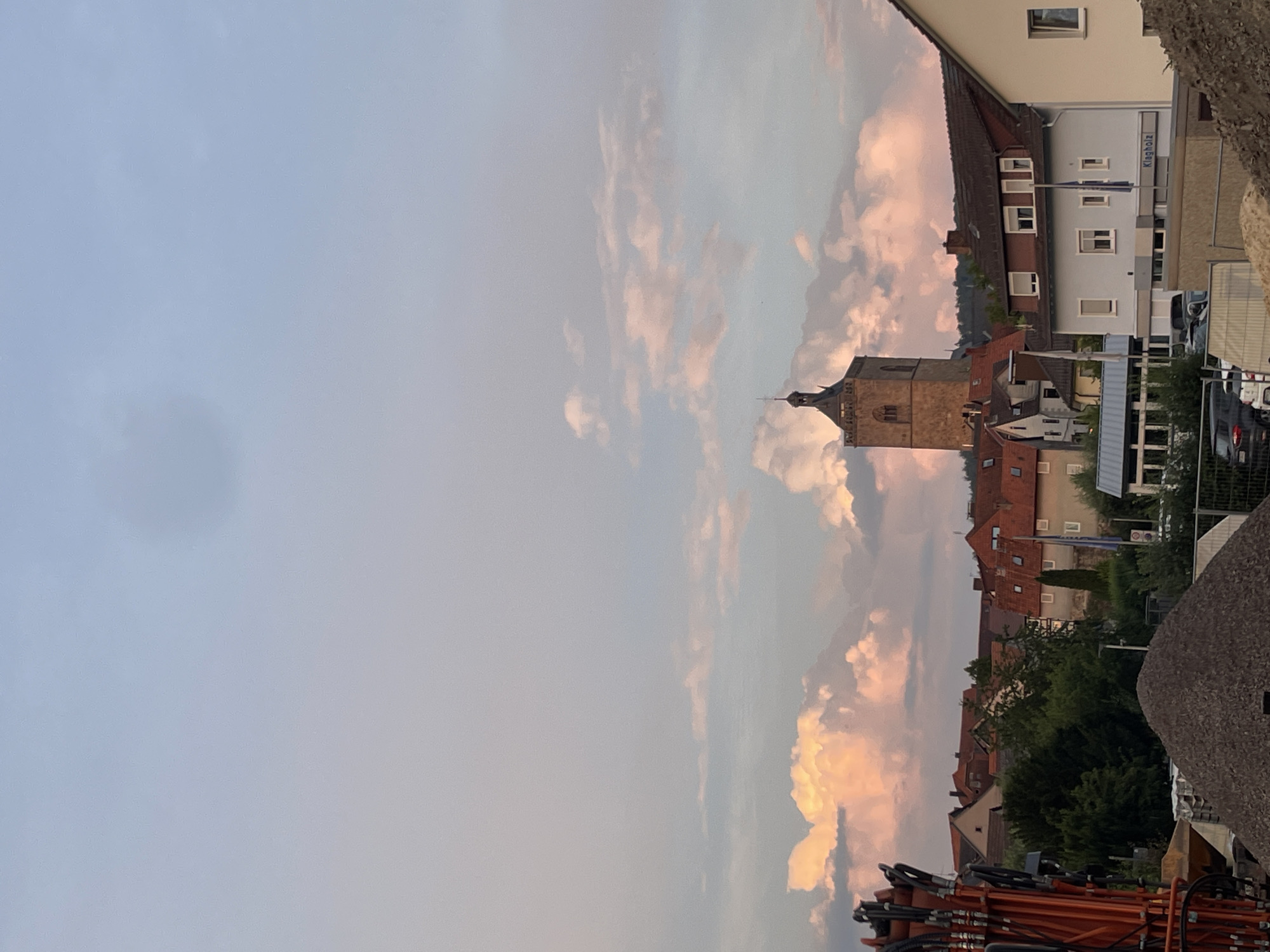 The image shows a view of the town of Neukirchen, Germany, with a prominent tower in the background and a construction site in the foreground. The sky is filled with dramatic clouds, tinged with pink and orange from the setting sun.
In the midground, a distinctive stone tower with a pointed roof and cross stands tall above a cluster of buildings with red-tiled roofs. The architecture suggests a historical or religious structure. The surrounding buildings are a mix of residential and commercial, with visible windows and dormers.
The foreground is dominated by a construction site. An orange excavator arm is visible on the left, and a large pile of gravel is in the center, partially obscuring the view of the town. A white van and several parked cars are visible behind a fence in front of a building with the sign "Klagholz". The overall impression is a blend of old and new, with a developing area set against a picturesque sky. There are no people visible in the image.