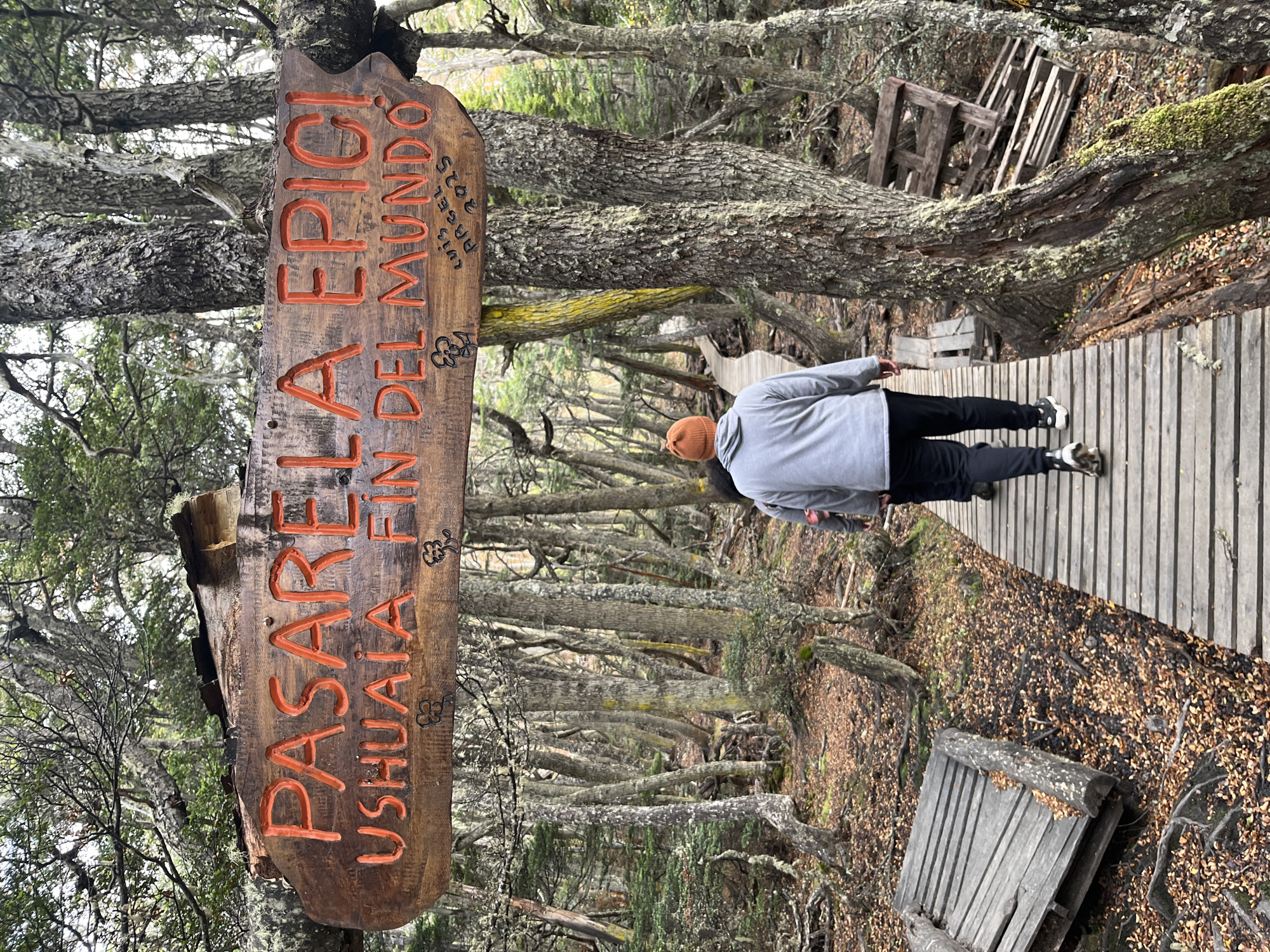 The image shows a wooden signpost that reads "PASARELA EPICA, USHUAIA FIN DEL MUNDO" in orange lettering. In smaller lettering it is marked "LUIS ABGEL 2025". A person wearing a light gray jacket, black pants, and an orange beanie is walking on a wooden boardwalk through a forest. The forest consists of bare trees with many branches. Fallen leaves cover the ground. There are wooden pallets placed along the side of the boardwalk. The sign indicates this is in Ushuaia, Argentina, known as the "end of the world."