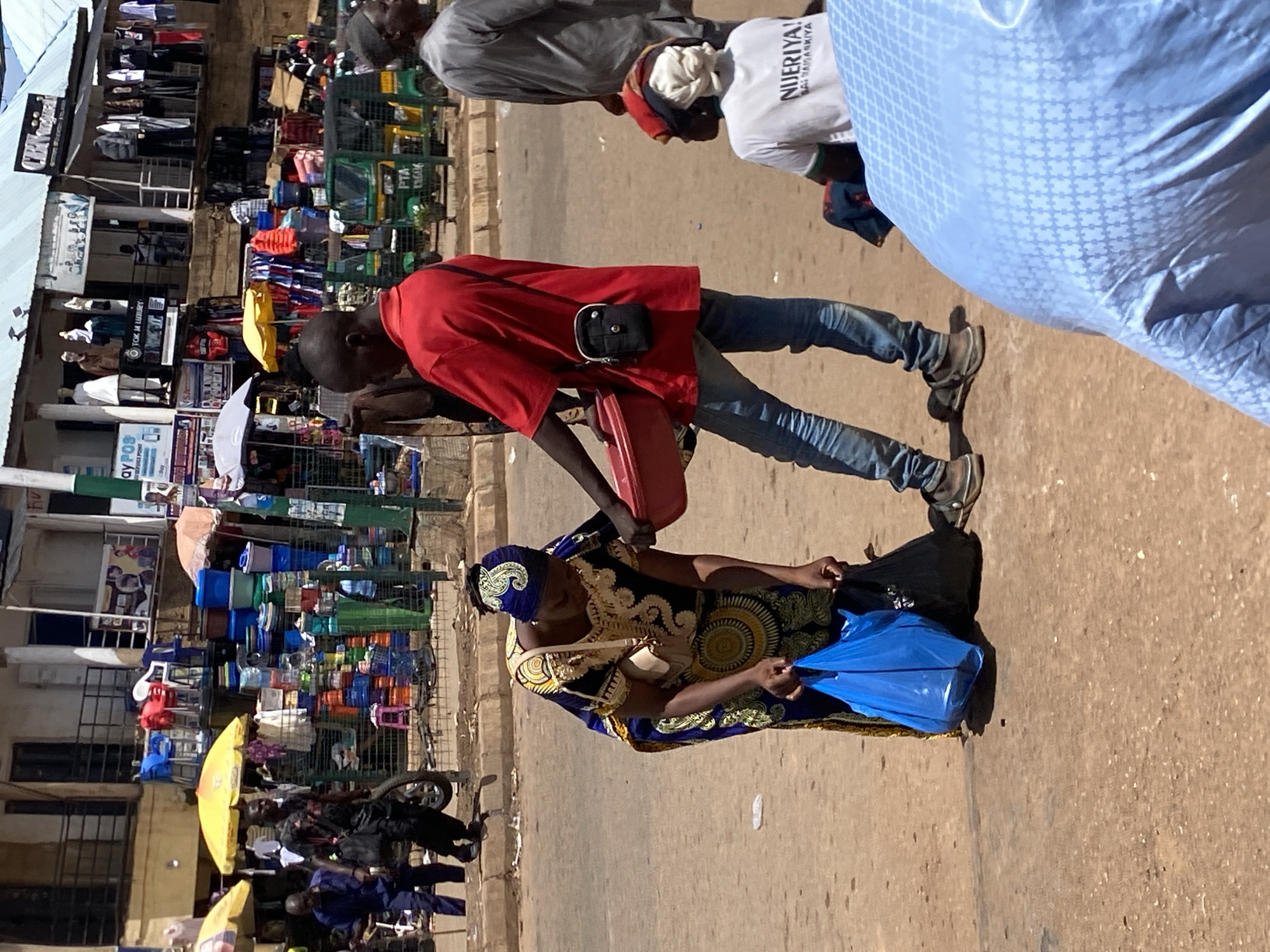 The image captures a vibrant outdoor market scene or busy street in Bukuru, Nigeria, under bright daylight conditions. The ground appears to be unpaved or dirt.
In the foreground, a man in a red short-sleeved shirt and blue jeans is prominently featured. He wears a small black pouch across his shoulder and holds a red, rectangular item, possibly a tray or container, in his hands. He is interacting with a woman who is either seated or crouched on the ground. This woman is dressed in a striking blue and gold patterned traditional outfit, possibly a boubou or wrapper, with a matching headscarf. She holds a bright blue plastic bag and a black bag beside her.
In the mid-ground and background, the scene is bustling with market activity. Various stalls and shops line the street, displaying a wide array of goods. One stall prominently features stacks of colorful plastic buckets, basins, and other household items. Other shops showcase clothing on hangers. Yellow market umbrellas provide shade to some vendors and areas.
Several other individuals are visible. A man in a light grey top is partially seen in the upper part of the image, and another person wears a white t-shirt with text. A green tricycle, commonly known as a Keke Napep, is also visible in the background.
Visible text includes:
* On the white t-shirt: "NIJERIYA! BALARABARBIYA" (likely "Nigeria! Our Nation" or similar slogan in Hausa).
* On the green tricycle: "PTA" and "1306A".
* On various shop signs: "OY POS", "TCM LUXURY", and a partially visible "OMAH".
The image conveys a sense of daily commerce and community interaction in a West African market setting.