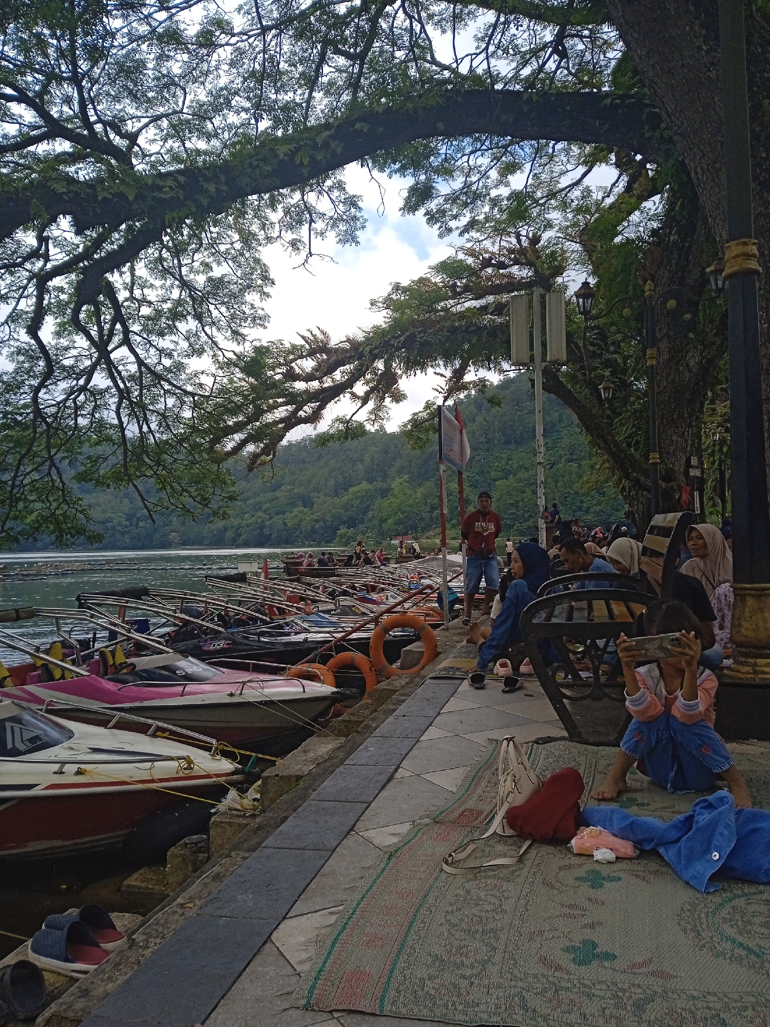 The image depicts a lakeside scene in Nglingi, Indonesia. Multiple motorboats and jet skis are docked along a stone walkway adjacent to the water. Trees overhang the walkway, providing shade. Several people are present; some are seated on benches or on a patterned mat, while others are standing near the water's edge. A young girl is seated on the mat, holding a smartphone in front of her face. Mountains are visible in the background. The environment suggests a recreational area or tourist spot.