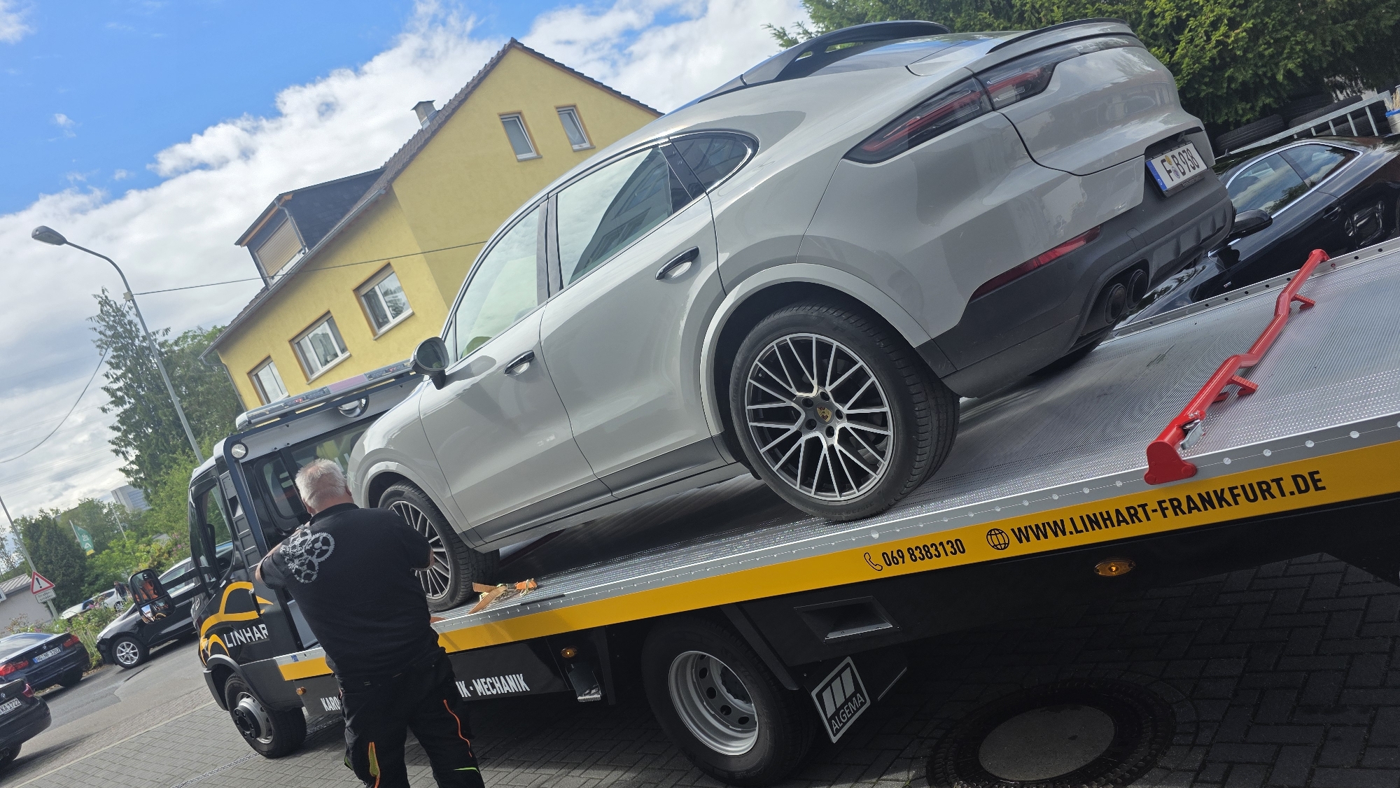 A silver Porsche Cayenne is being loaded onto a tow truck with a flatbed. The car is positioned diagonally on the truck, with its rear on the flatbed and its front angled upwards. A tow truck driver, wearing a black shirt with a gear logo and black pants with orange stripes, is standing by the truck, looking at the car. The tow truck has "LINHAR" written on its side, along with a phone number and website address. In the background, there are yellow houses, trees, and a blue sky with clouds. Other cars are visible parked on the street.