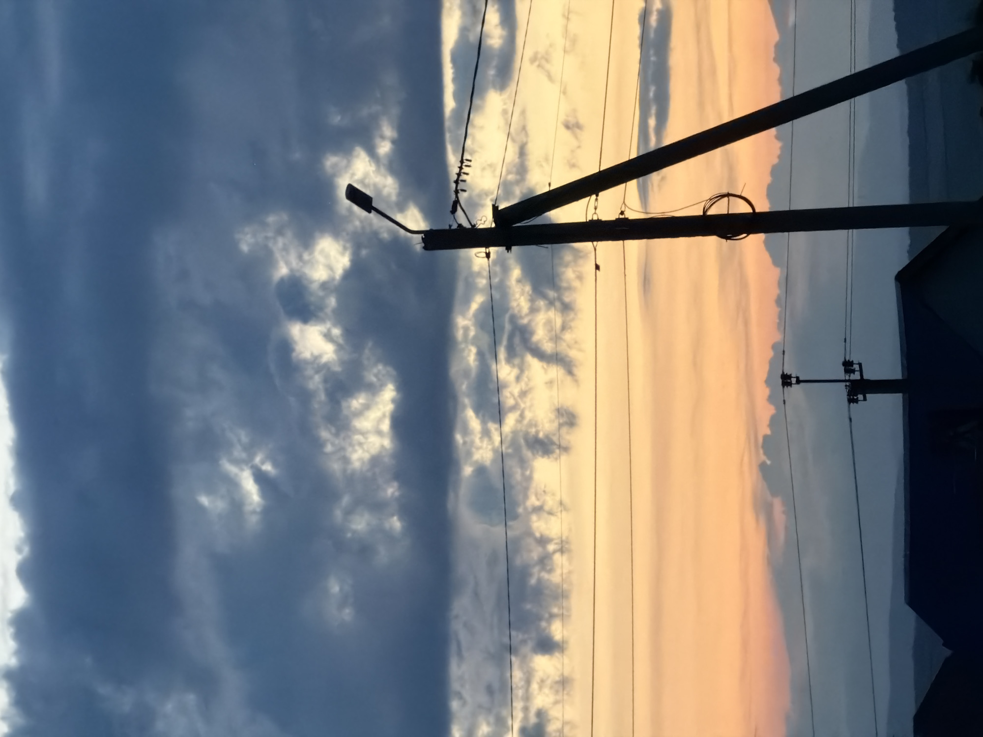 This media file shows a vertical outdoor scene captured during dusk. The sky is split into two distinct halves. The left side is dominated by dark, dramatic storm clouds, while the right side is a softer expanse of peach and orange hues, indicative of a sunset. Power lines and utility poles are prominently featured throughout the frame, silhouetted against the sky. A street light is visible on a pole in the left portion of the image. In the far right, the edge of a dark structure, possibly a building, is discernible. The overall mood is serene yet slightly ominous due to the contrast between the stormy and clear sky. No text is visible in the image.