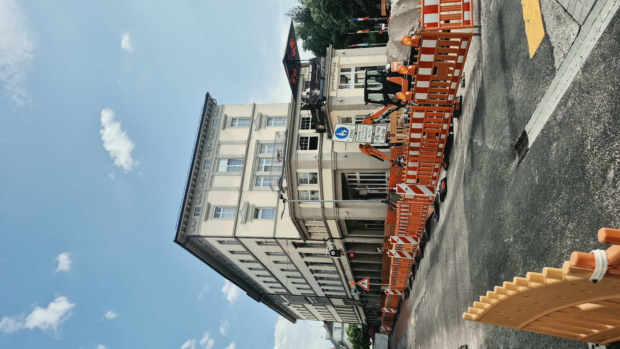 The image captures a street scene in Baden-Baden, Germany, with ongoing construction work. A multi-story building with a light-colored facade dominates the right side of the frame. The building features multiple windows, balconies, and architectural details. A sign for "PORTERHOUSE GRILL" is visible on the building's facade, along with an awning and flags.
The street in front of the building is partially closed off with orange and white construction barriers. An excavator is present on the site, positioned behind the barriers, next to a pile of sand. The road surface is asphalt, with some markings and a grate visible. The sky is blue with scattered white clouds. The overall atmosphere suggests a commercial area undergoing renovations or street repairs. There are no people clearly visible in the image, but there are indications of activity related to the construction.