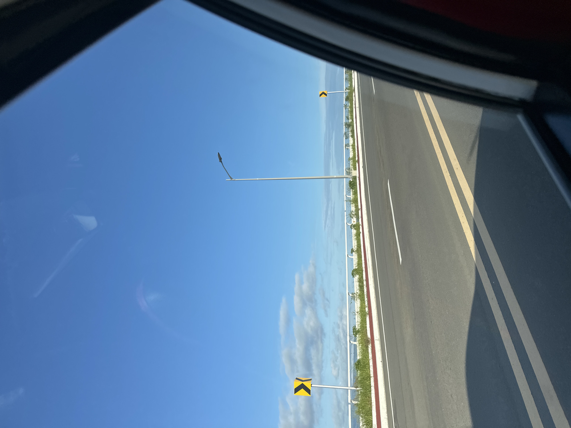 The image displays a daytime view from inside a vehicle, looking out the passenger side window onto a two-lane asphalt road. The vehicle's dark window frame and a red interior element are visible along the right edge. A prominent shadow, presumably from the vehicle itself, extends across the lower left portion of the road.
The asphalt road features double solid yellow lines separating the lanes and a solid white line delineating the right edge of the pavement. To the left of the road, a concrete curb with a red painted top borders a strip of green grass and sparse vegetation. Beyond this, a white metal guardrail runs parallel to the road.
A tall, grey metallic streetlight pole with a single lamp fixture is positioned on the left side of the road in the mid-ground. Two yellow chevron signs are visible, both indicating a left curve. One is closer to the foreground on the left side, mounted on a grey pole. The second is visible further down the road on the right side, partially obscured by the vehicle's frame.
Beyond the guardrail, a calm body of blue water, consistent with a sea or ocean, stretches to the horizon. Above, a bright, clear blue sky dominates the upper portion of the image, dotted with scattered white cumulus clouds. No individuals are present in the scene. This appears to be a coastal road in a sunny environment, located in Davao City, Philippines.